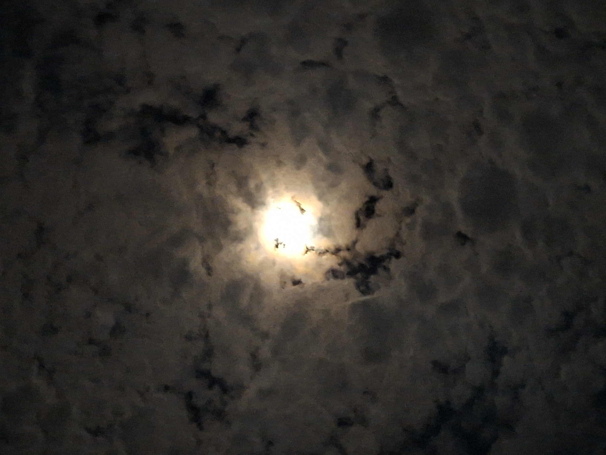 The image shows a full moon shining through scattered clouds at night. The moon is bright and slightly obscured by the clouds, creating a hazy effect. The clouds are dark and textured, appearing to be thin and wispy in some areas and more dense in others. The overall atmosphere of the image is dark and moody. There is no indication of the city of Kruengmate, Indonesia, or any other specific location. The image is a simple night sky photograph with no discernable people, objects, or actions.