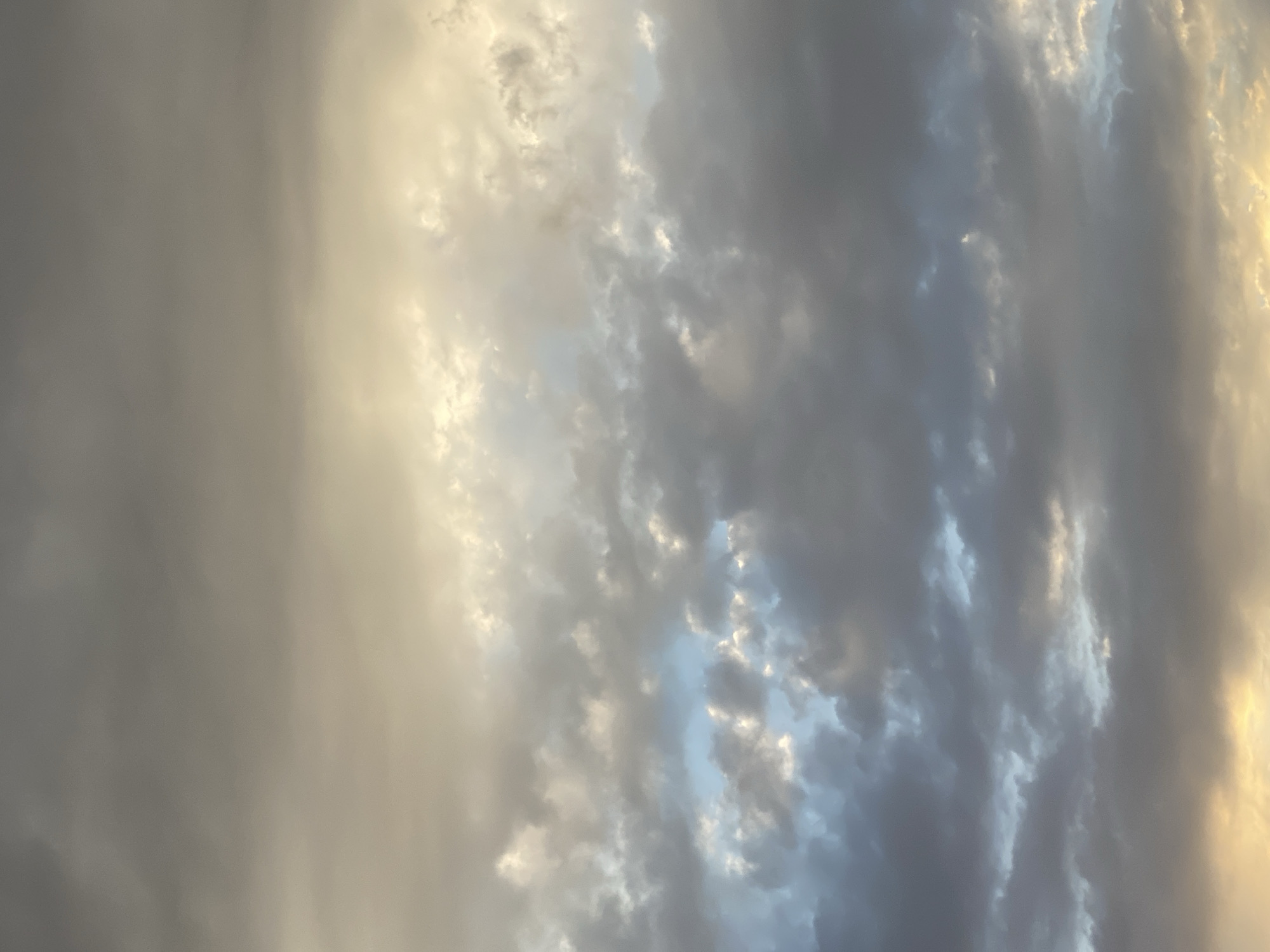 This is a photograph of a cloudy sky. The clouds are dark and grey in some areas, and lighter and yellowish-white in others. There are patches of bright blue sky visible between the clouds. The overall impression is of a dynamic sky, possibly on the verge of rain or clearing. The image is taken from a low angle, looking upwards. There are no people, objects, or man-made structures visible, only the sky and clouds. The location in Bishkek City, Kyrgyzstan, is not discernible from the image itself.