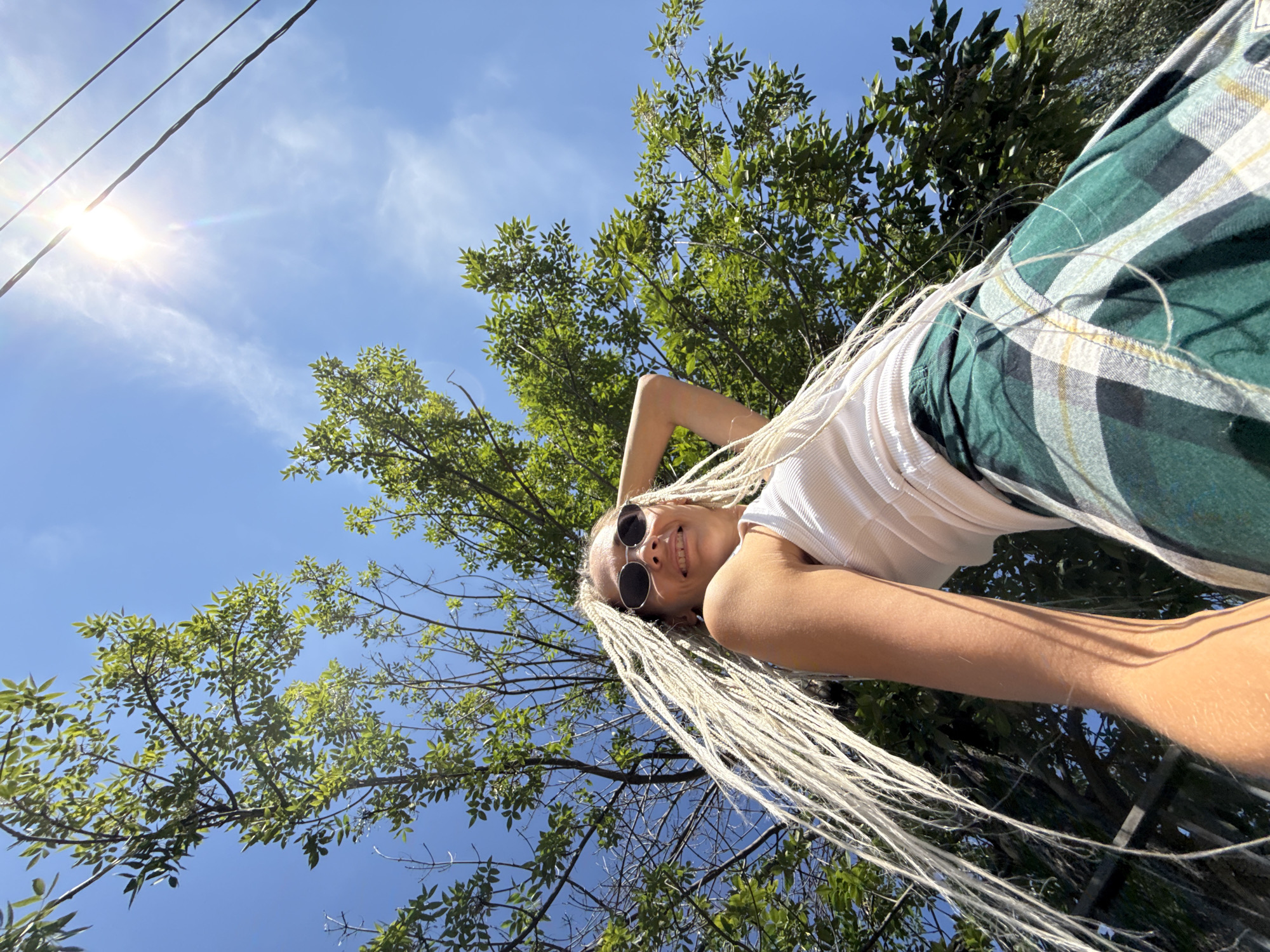 A low-angle, medium shot captures a young woman with blonde braided hair from above, looking up towards the sun. She wears sunglasses, a white tank top, and a green plaid skirt. Her right arm is raised, touching her head. Lush green foliage from a tree frames the upper left of the image, while power lines and the bright sun are visible in the upper right. The sky is a clear, vibrant blue. The scene appears to be outdoors. The specific location in the city of Rusanovo, Russia, cannot be determined from the visual information provided.