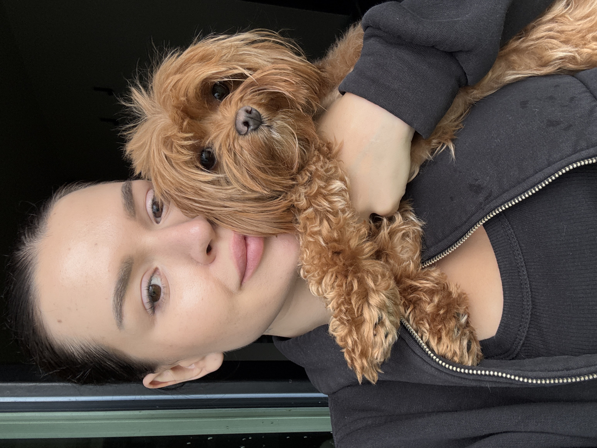 This is a close-up, vertically oriented selfie of a young woman with a small, reddish-brown dog. The woman's face is partially obscured by the dog, which is being held close to her neck and chest. She is wearing a black hooded zip-up jacket. The dog appears to be a Cavapoo or a similar breed with curly fur. Its head is resting on the woman's shoulder, and its paws are visible on her chest.
The woman's skin is clear and her makeup appears minimal. Her eyes are open, and she is looking towards the camera. The dog has dark eyes and a dark nose. The background is dark and out of focus, but appears to be indoors. The lighting suggests it could be daytime, possibly overcast, given the diffused light on the woman's face. There are no discernible external location cues, other than the provided context of Prague, Czechia, and no visible text. The overall impression is one of affection and comfort between the woman and her dog.