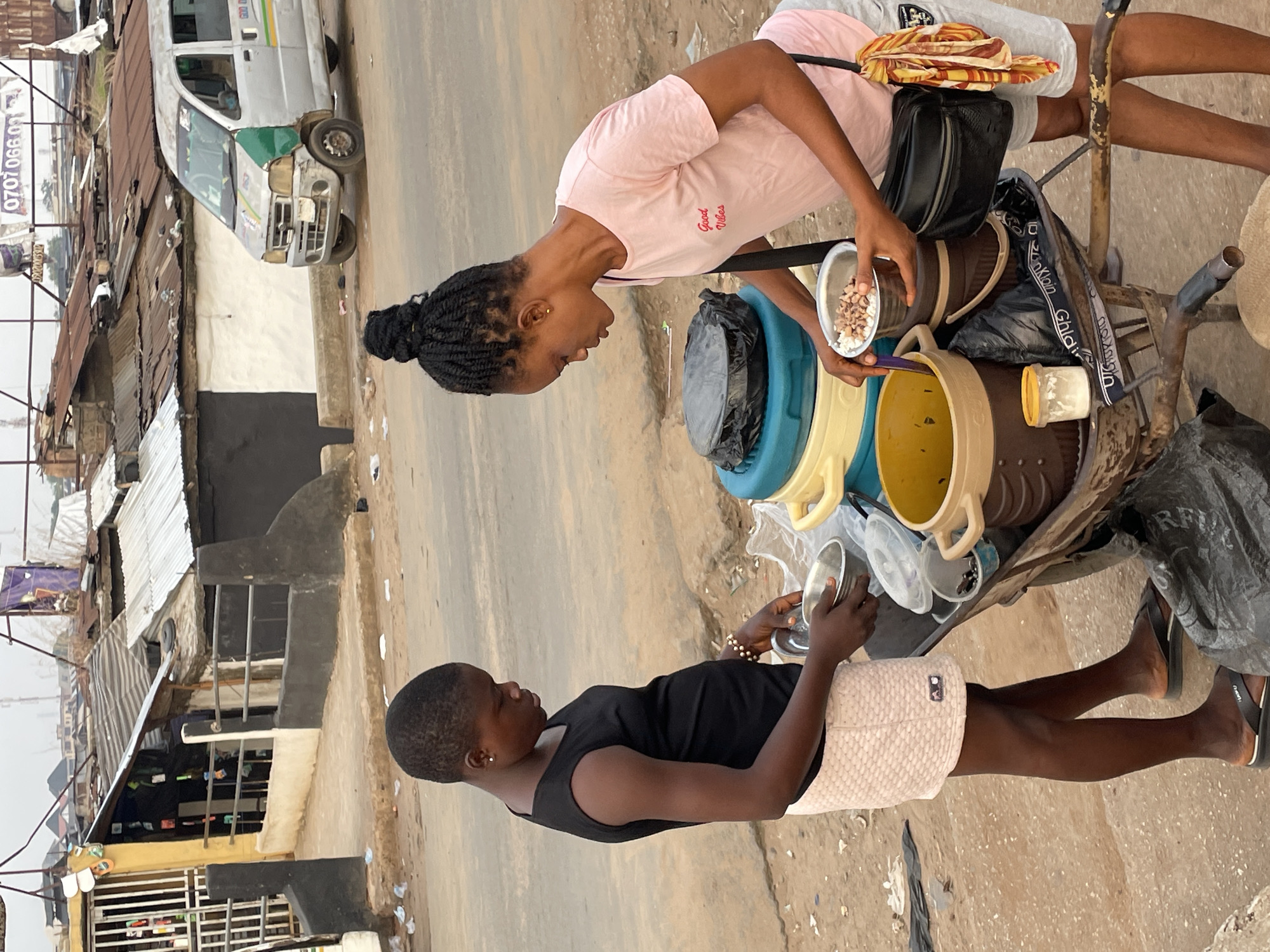 Two young women stand beside a cart. The woman on the right wears a pink t-shirt with the words "Good Vibes" on it and grey shorts. The woman on the left wears a black top and beige shorts. The cart holds several containers, including a blue and yellow plastic container. The woman in pink appears to be serving the woman in black something from a bowl. In the background, there are buildings, one with a black and white awning, and a white van. The image appears to be taken on a street in Onitsha, Nigeria.