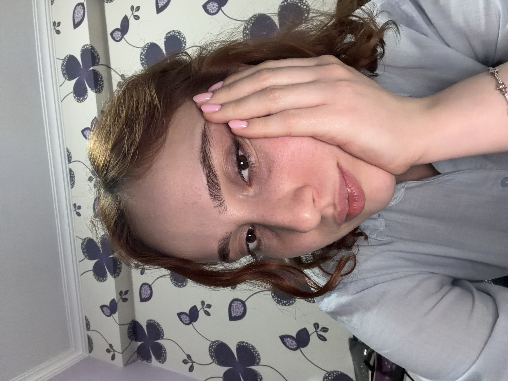 A young woman with auburn hair is close to the camera, partially obscuring her face with her left hand. She wears a light blue collared shirt and a star-shaped bracelet on her wrist. The background features a white wall with a repeating pattern of stylized purple flowers and green leaves. A white-framed door or molding is visible on the left. The image does not provide enough information to determine the location in Tbilisi, Georgia.