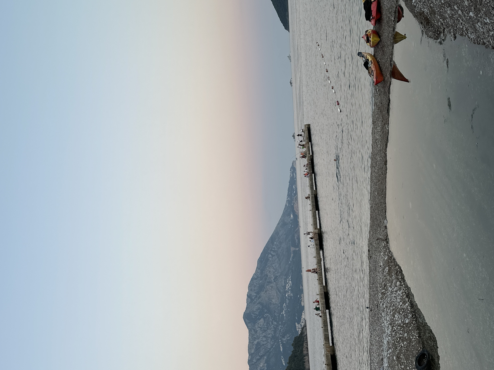 The image depicts a serene evening scene along the coast of Budva, Montenegro. In the foreground, a gravelly beach area is partially covered by a calm, reflective body of water. Three kayaks, red, yellow, and orange, are resting on the beach near the water's edge. Further back, a long, concrete pier extends into the calm sea. Several people are visible on the pier, some seated and others standing, appearing to be enjoying the view or fishing. A lone swimmer is in the water near the pier. In the background, a majestic, rugged mountain range dominates the horizon, with buildings nestled on its lower slopes. The sky above transitions from a soft pink near the horizon to a pale blue, indicating dusk. Several boats are scattered across the distant sea, adding to the tranquil maritime atmosphere.