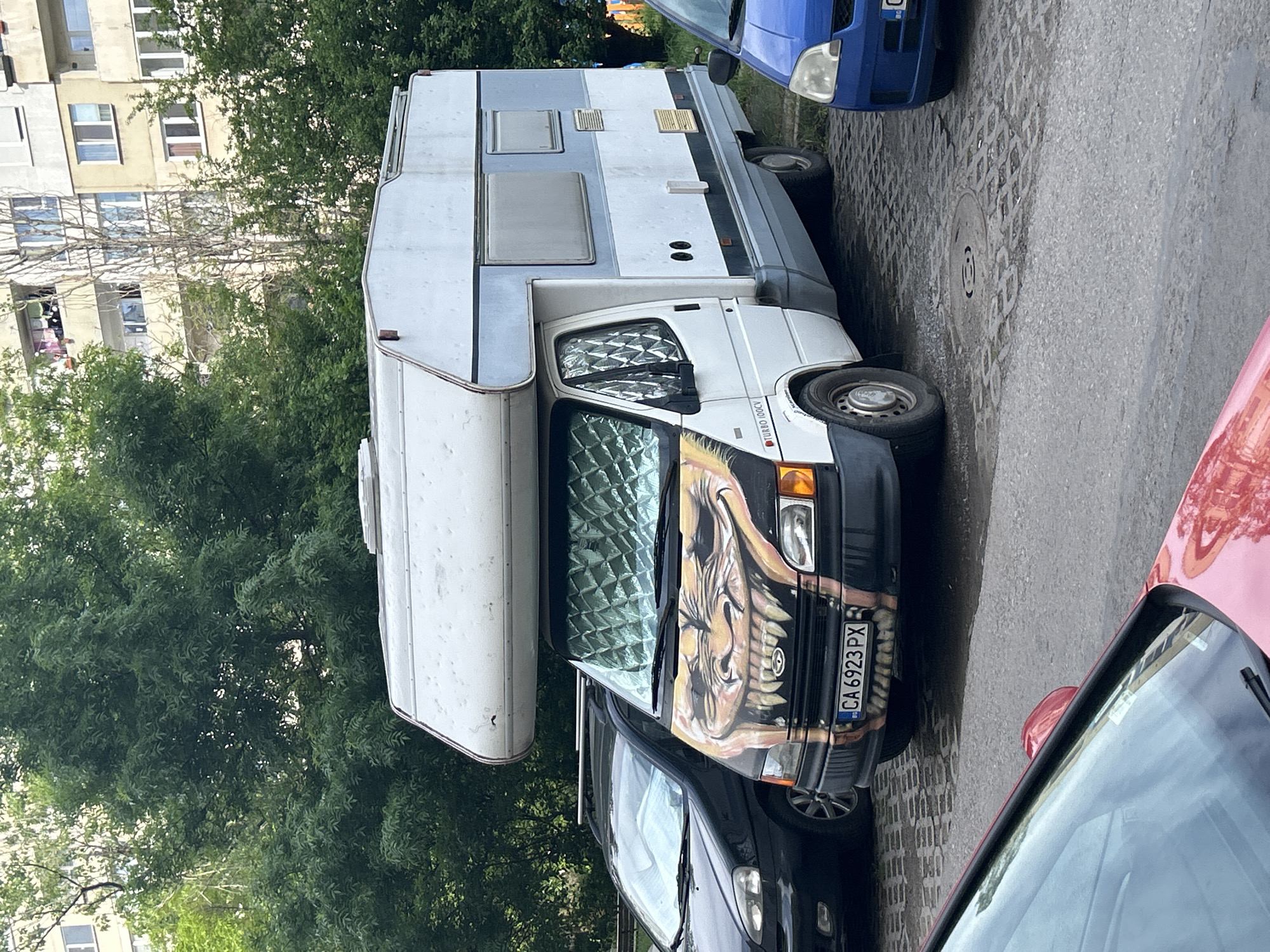 The image shows a white camper van parked on a city street, with the Bulgarian license plate "CA 6923 PX". The van has a distinctive painting of a monster face on the hood. The van is sandwiched between a dark-colored car and a red car. To the left is a tree, and behind it is an apartment building. The location appears to be in a residential area.
