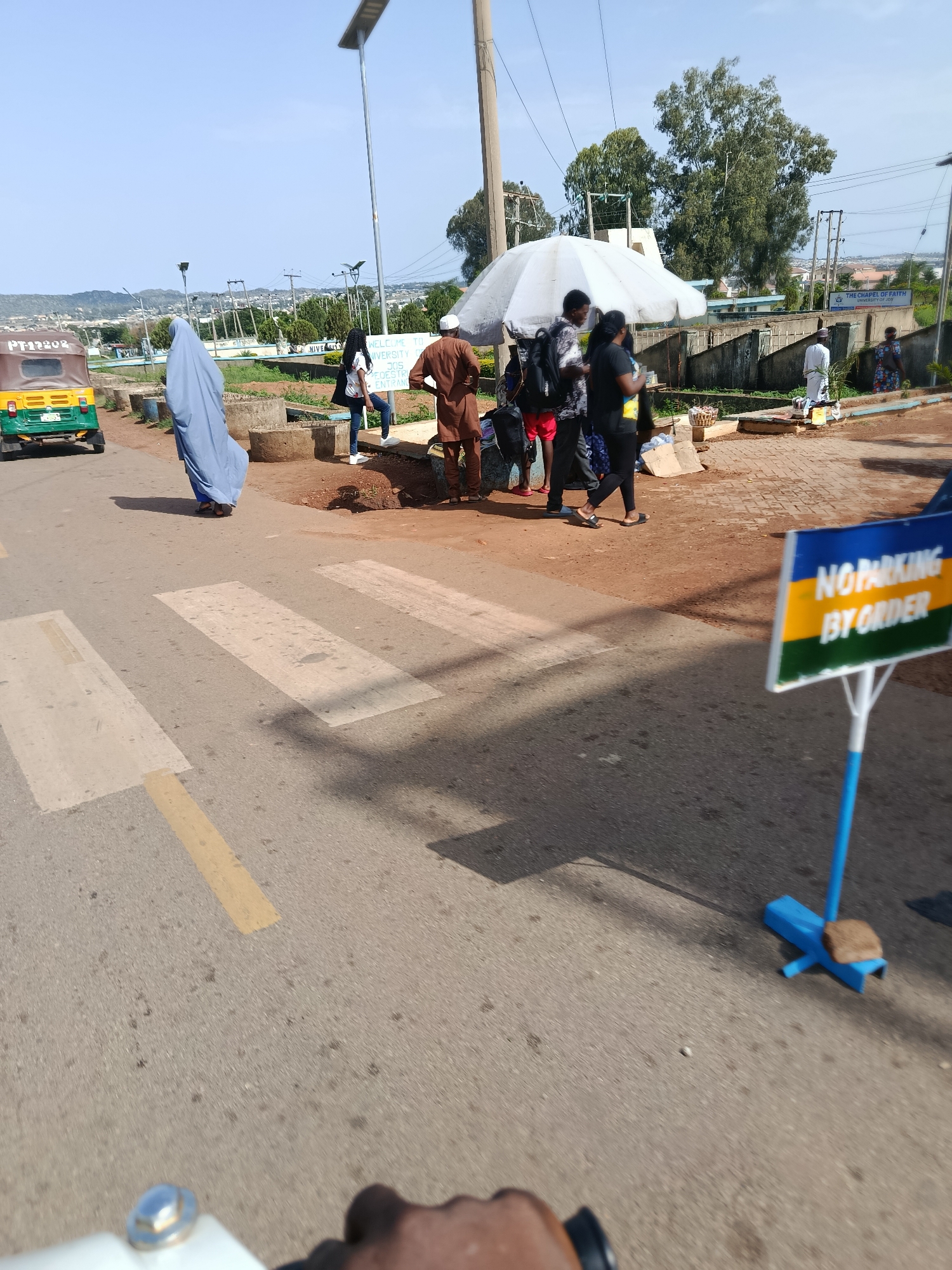 This image captures a scene on a street in Jos, Nigeria, specifically near the University of Jos. In the foreground, a pedestrian crossing is visible on an asphalt road. To the left, a yellow and green auto-rickshaw is parked. A person in a light blue abaya stands near the pedestrian crossing, with their back to the camera. Further back, a group of people are gathered under a white umbrella, some appearing to be waiting or interacting. One individual is seen wearing a brown traditional outfit, and another in a white kaftan. Behind the group, a wall with signage can be observed, indicating "THE CHAPEL OF FAITH UNIVERSITY OF JOS". A blue and yellow "NO PARKING BY ORDER" sign is placed prominently in the foreground on the right side of the image. The background shows a cityscape with buildings, trees, and a clear blue sky, suggesting a sunny day. The overall impression is one of a bustling street scene with people engaged in various activities near a prominent university landmark.