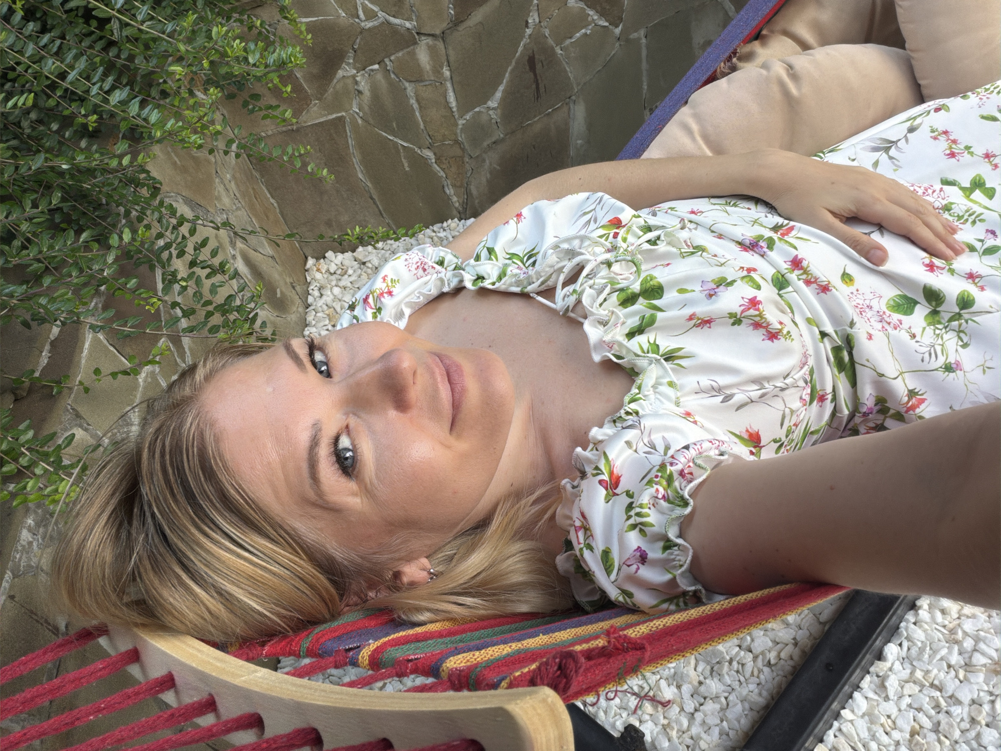 A Caucasian woman with blonde hair pulled into a bun, wearing a white dress with a floral print, lies in a hammock. She is looking at the camera and smiling. The hammock is made of colorful fabric and is suspended by wooden beams. In the background, there is a stone wall and green foliage. The scene appears to be outdoors, possibly in a garden or patio area. The location is specified as Nizhnyaya Shilovka, Russia.