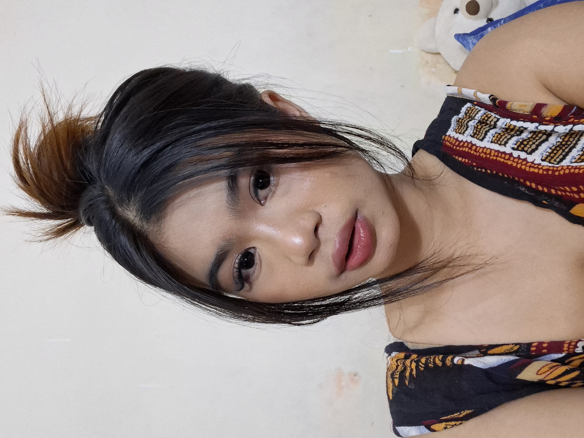 A close-up, head-and-shoulders photograph shows a young woman with dark hair styled in a messy bun with some strands falling forward. She has prominent eyelashes, dark eyebrows, and glossy, pink lips. Her skin has a dewy sheen. She is wearing a sleeveless top with a patterned design featuring abstract shapes in black, yellow, orange, red, and white. The background is a plain, light-colored wall with a slight discoloration. To the right of the woman, a white stuffed bear is partially visible. The setting appears to be an indoor environment, likely a bedroom, as suggested by the presence of the stuffed toy and the casual attire. There is no indication of the specific location within Kuta, Indonesia.