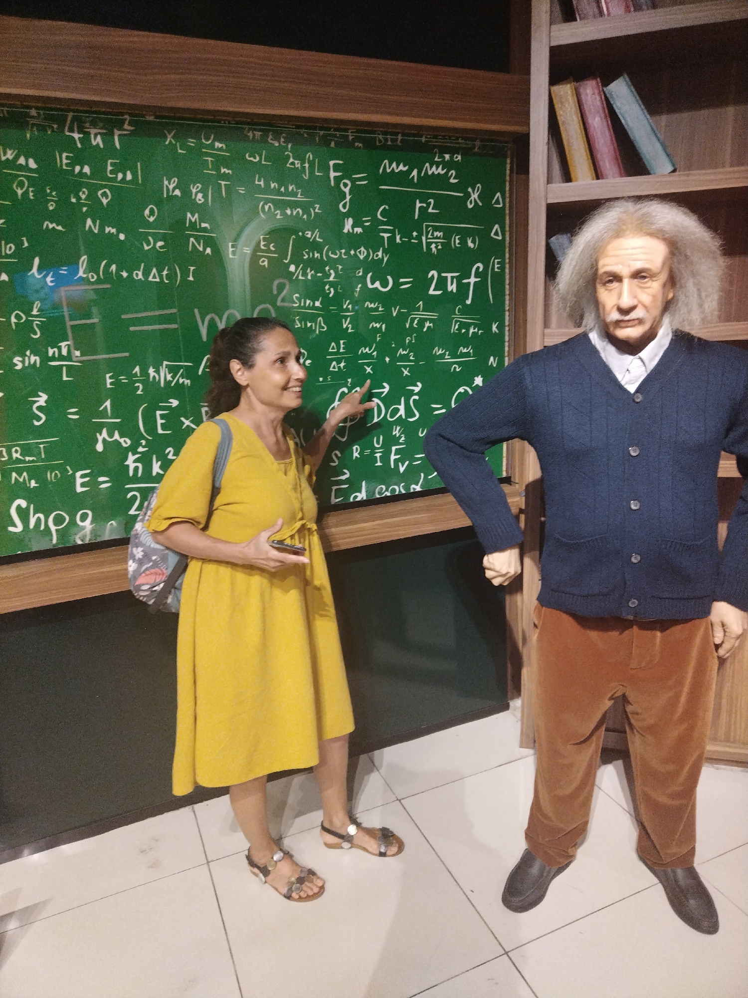 A medium shot captures a woman in a yellow dress standing next to a wax figure of Albert Einstein in an indoor setting. The woman is turned towards the wax figure, her right hand pointing towards the chalkboard behind Einstein, which is covered with mathematical equations. She is holding a mobile phone in her left hand.
The Einstein figure is dressed in a dark blue cardigan over a white collared shirt, paired with brown trousers and black shoes. Behind him is a wooden structure that resembles a bookcase filled with books.
The background is a chalkboard full of complex equations. The floor is tiled in light-colored squares. The lighting is bright and artificial, suggesting the scene is indoors, possibly in a wax museum located in Unknown, Unknown.