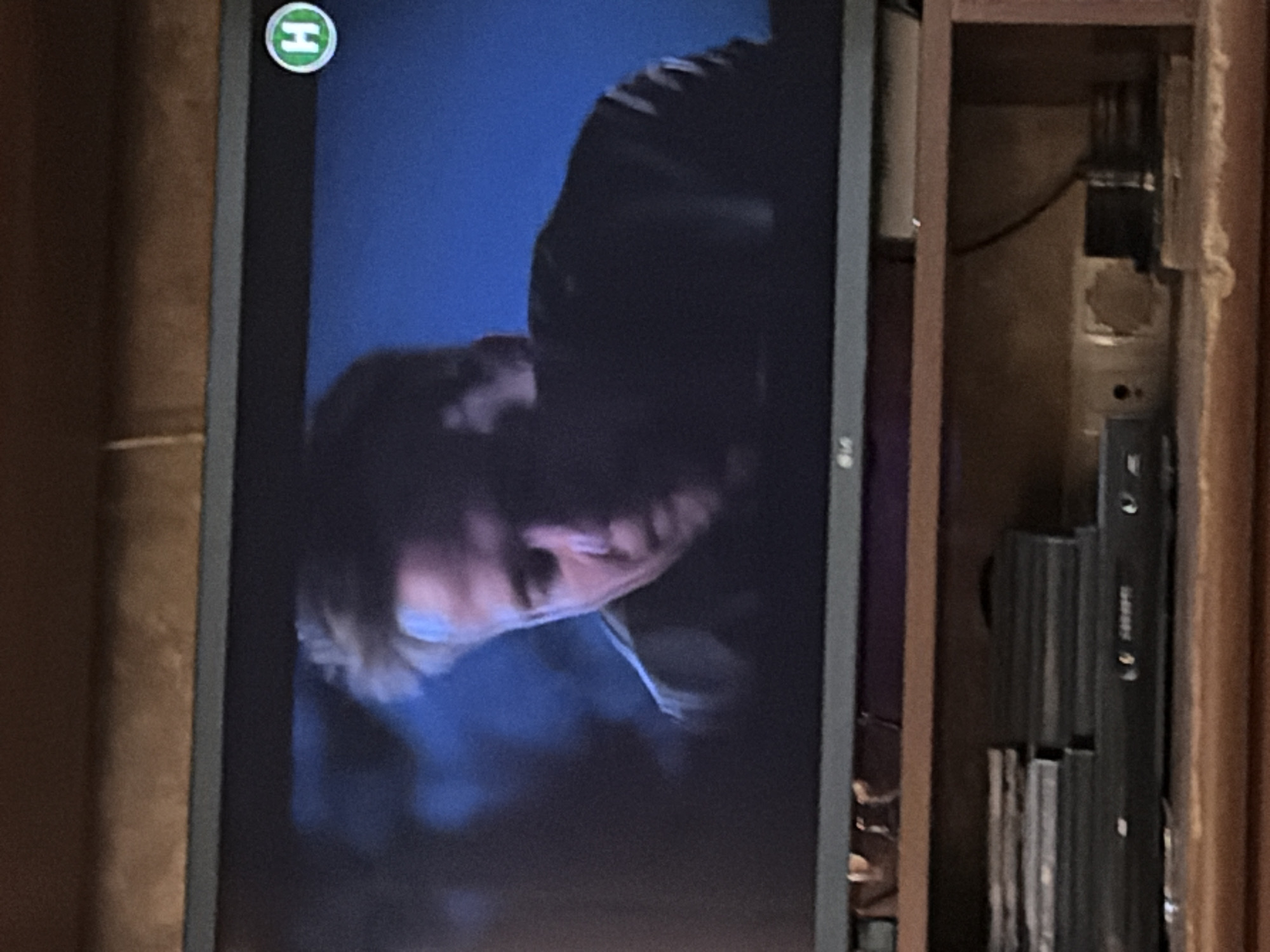 A television screen displays a close-up of a man's face with light-colored hair, looking down and to the right. He is wearing a dark jacket. A circular green logo with a white "H" is visible in the upper right corner of the screen. The background of the screen is dark blue. Below the television, a wooden shelf holds stacks of DVDs and a DVD player. The lighting suggests an indoor setting at night.