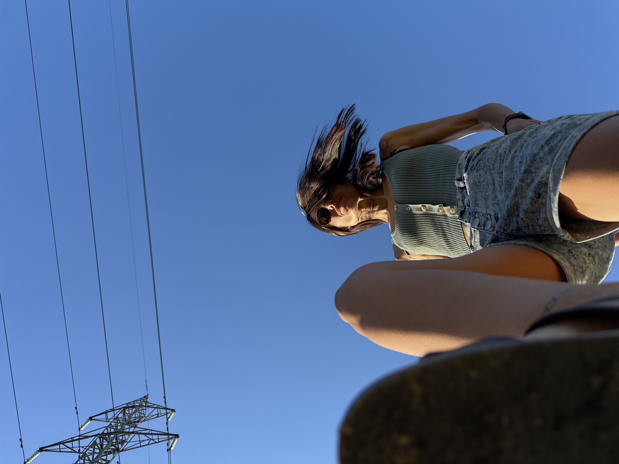 From a low angle, the image depicts a woman with dark hair and sunglasses, wearing a green tank top and denim shorts. She is positioned above the camera, her hair blowing in the wind. An electrical transmission tower and power lines are visible against a clear blue sky. The image was taken in Pleven, Bulgaria.