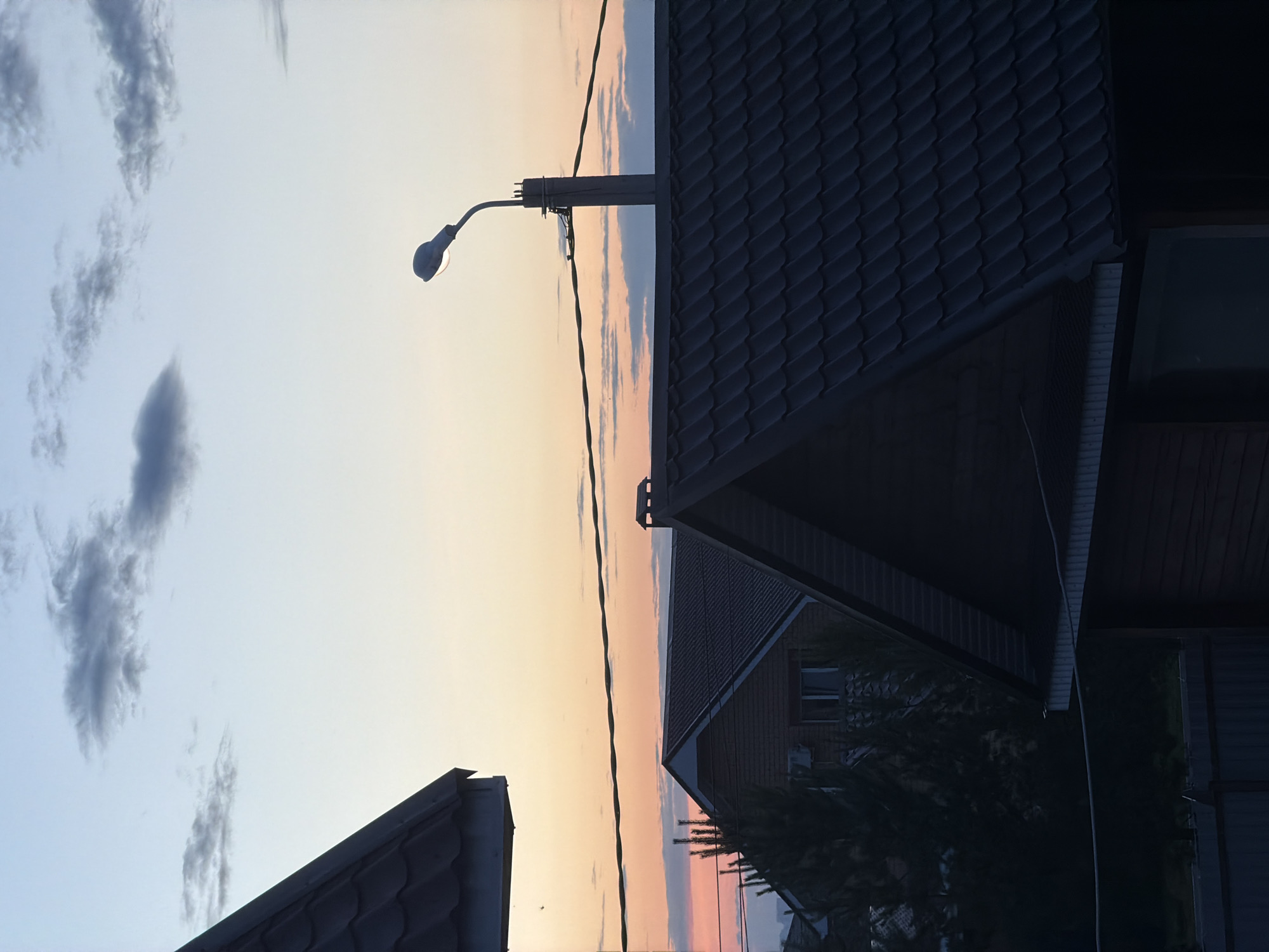 The image depicts a twilight scene over a residential area, possibly in the village of Shmid tovo, Russia. In the foreground, the dark, tiled roofs of several houses dominate the lower half of the frame. The roof on the far left is partially visible, angled towards the top left corner. Another prominent roof occupies the center-right, with a sharp gable. A smaller section of a wooden structure with horizontal siding is visible at the bottom center.
Above the rooftops, power lines stretch horizontally across the scene. A street lamp on a pole is visible on the right side, its light illuminated, piercing the fading light. The sky displays a gradient of colors, transitioning from a pale orange and pink near the horizon, indicative of a sunset, to a soft blue at the top, with scattered, wispy clouds. A few dark clouds are present in the upper portion of the sky, contrasting with the lighter hues. Trees are visible behind the houses, appearing dark and silhouetted against the lighter sky. No people are visible in the image.