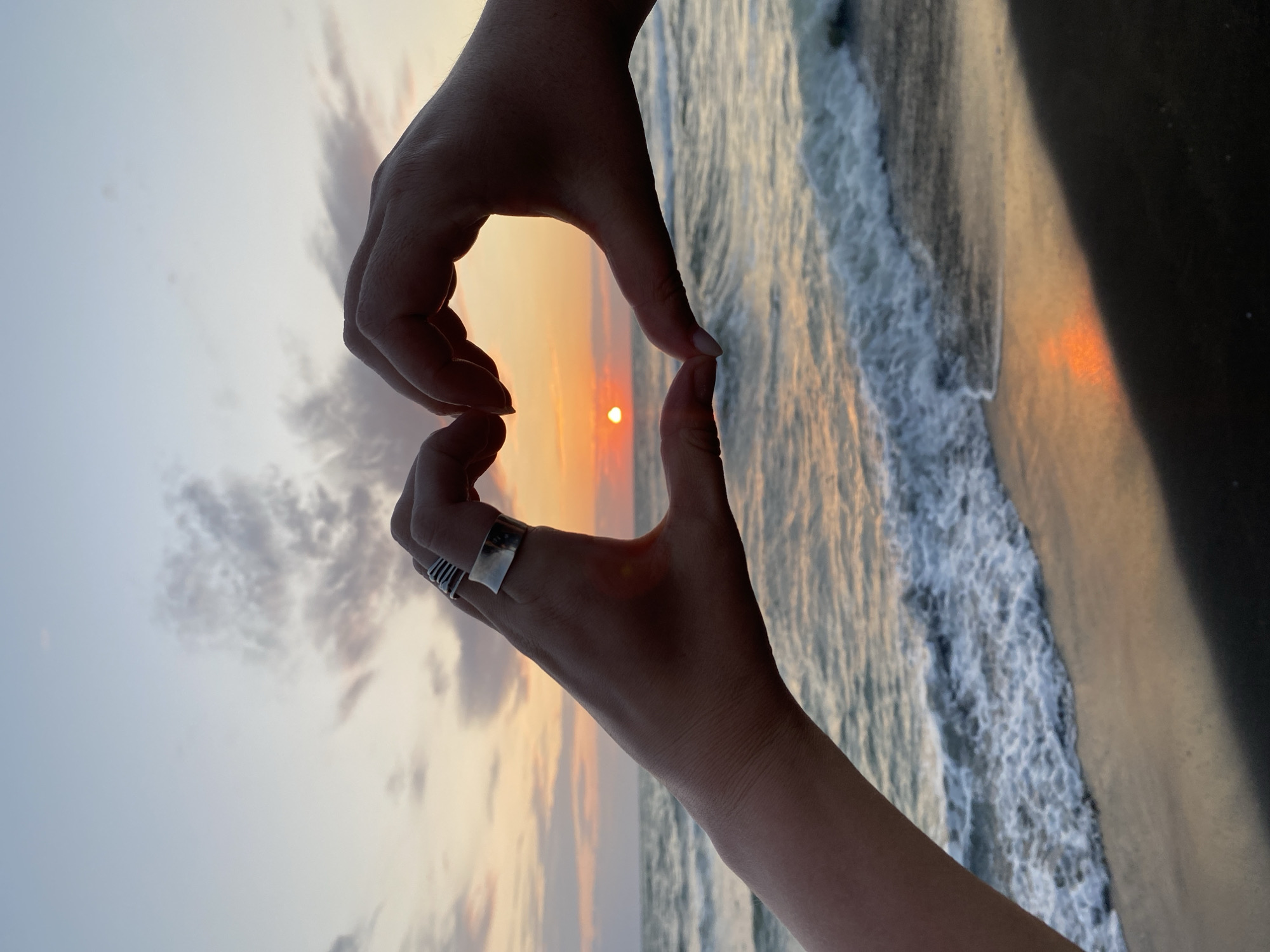 Two human hands form a heart shape in the foreground, framing a sunset over the Black Sea in Ureki, Georgia. The sun appears as a small, bright orange orb near the horizon, casting a warm glow on the sky and the water. The ocean exhibits gentle waves breaking on the shore, and the wet sand reflects the colors of the sunset. The sky above is a gradient of pale blue and soft pinks, with scattered clouds. One hand is adorned with several rings. The scene conveys a sense of romance and tranquility.