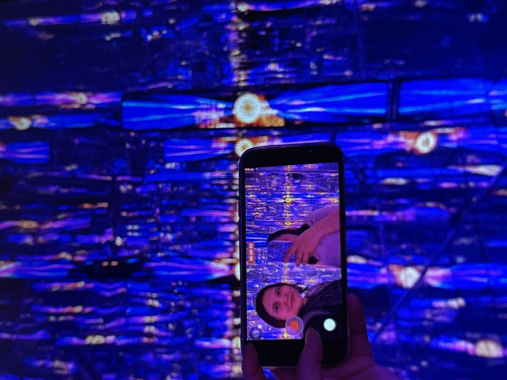 A person is holding a smartphone to take a selfie in a room with blue and purple lights. The smartphone screen shows two women taking a selfie, and the background on the screen is a reflection of the lights. The main background of the image is a blurry view of the light installation.