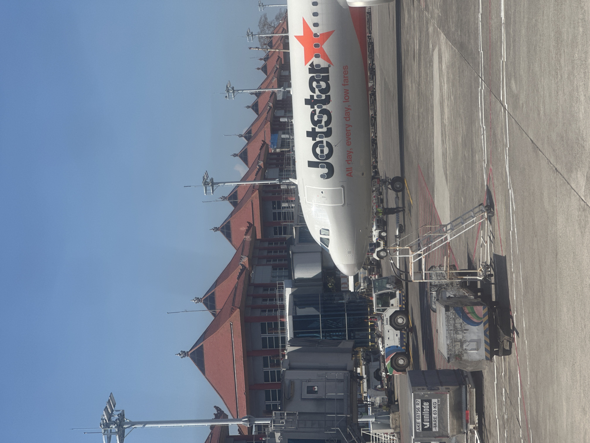 An airplane with "Jetstar" written on its side is parked at an airport gate in Kuta, Indonesia. The airplane's nose is visible, with the cockpit windows looking out onto a bright, clear sky. The airplane is connected to a jet bridge, which leads to a modern airport terminal building with a distinct red-tiled roof and traditional Indonesian architectural elements. In the foreground, several airport service vehicles and baggage carts are visible on the tarmac. A few airport personnel can be seen near the vehicles. The scene appears to be busy with airport operations.