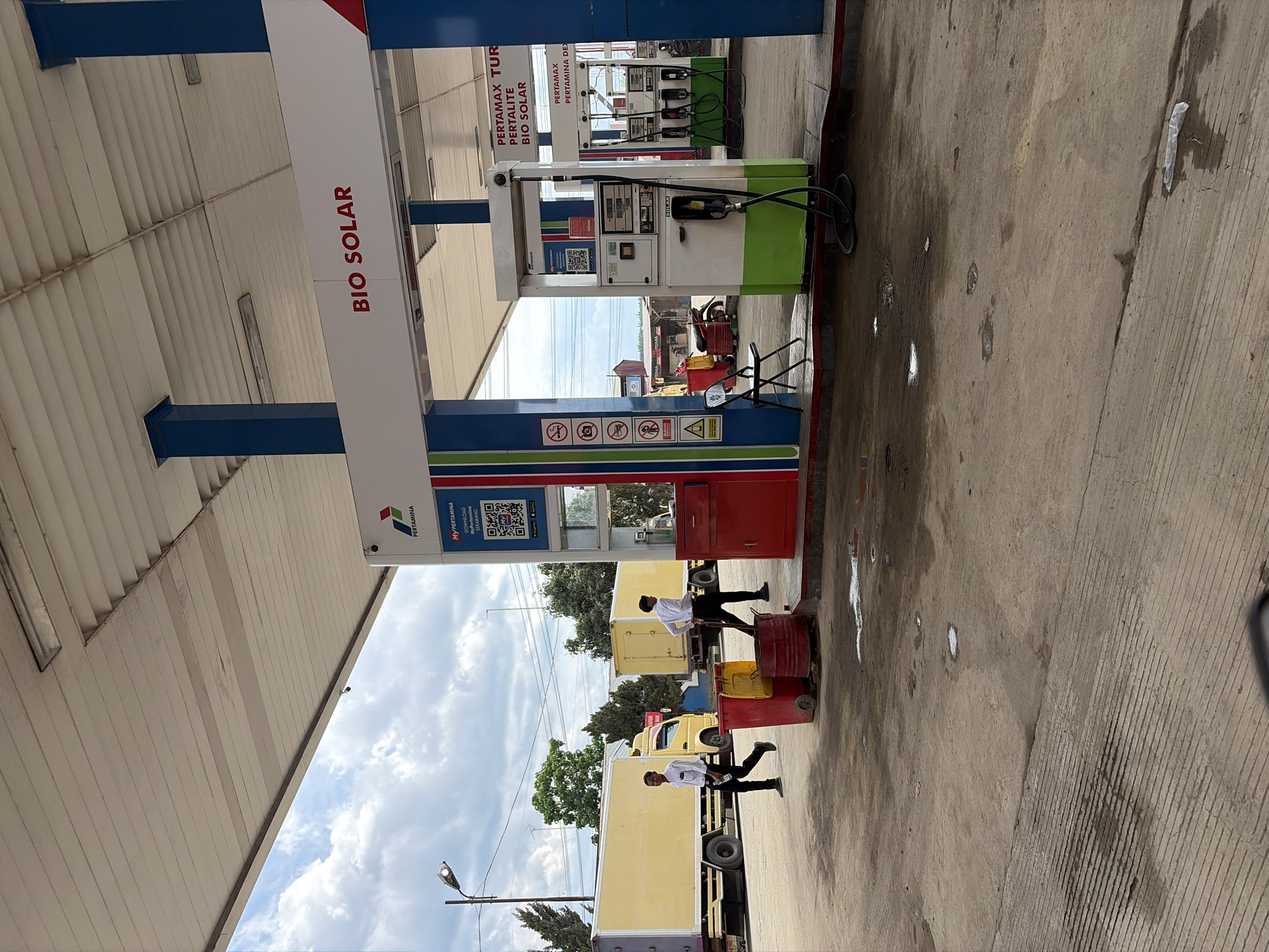 The image shows a Pertamina gas station in Simpangempat, Indonesia. The gas station has pumps offering Bio Solar, Pertamax Turbo, Pertalite, Bio Solar, Pertamax, and Pertamina Dex. Two individuals in white shirts and dark pants appear to be moving red and yellow barrels with wheels. There are light yellow trucks parked nearby. The sky is cloudy. The gas station canopy is white with blue supports. The ground is concrete and shows signs of wear.
