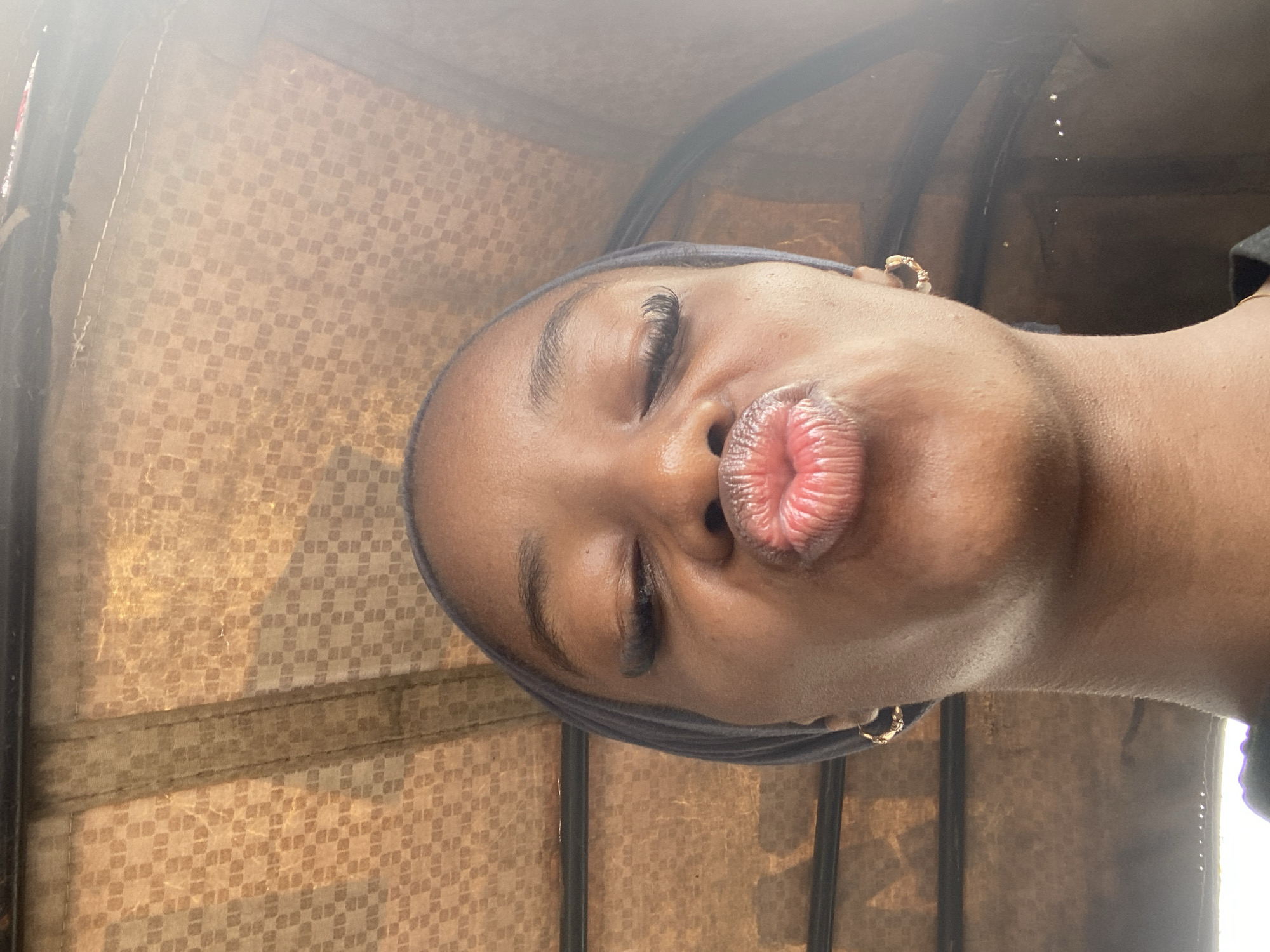 The image shows a close-up, front-facing shot of a young Black woman with her eyes closed and lips puckered as if to give a kiss. She is wearing a dark headscarf and small hoop earrings. The background is a textured, brownish material, possibly the interior of a vehicle, with a faint checkered pattern. Sunlight is casting a glow on the top right of the image, creating a bright, hazy effect. There is no indication of the city of Jos, Nigeria, in the image.