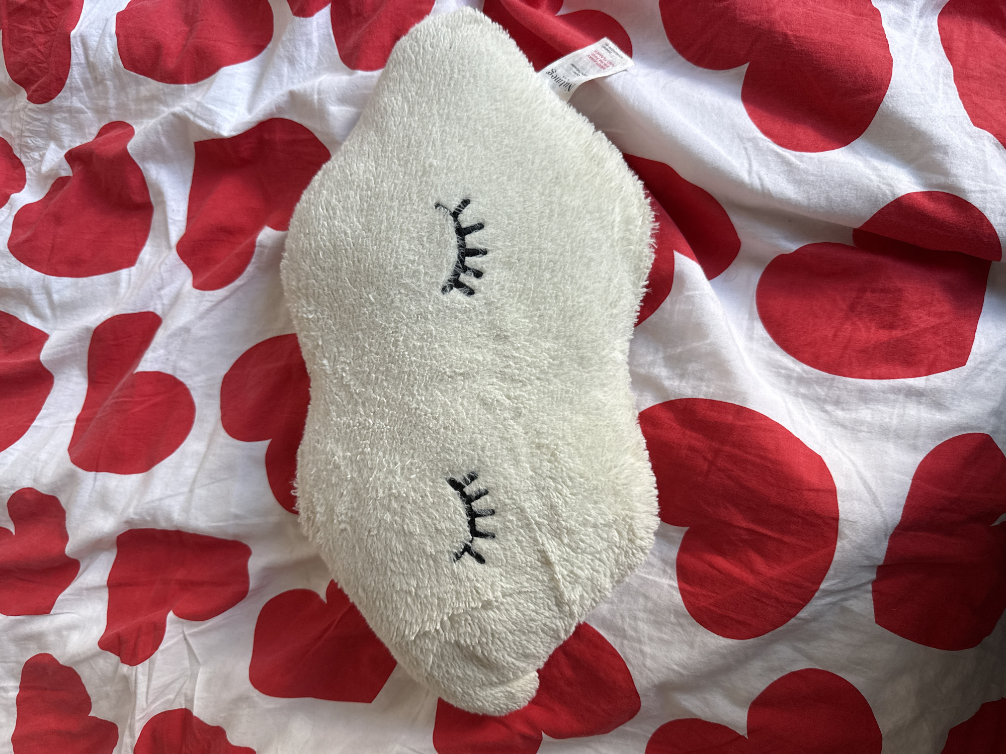 An overhead shot captures a plush, white, cloud-shaped pillow with embroidered closed eyes, resting on a white fabric background adorned with a pattern of large red hearts. The pillow is slightly off-center to the left. The fabric appears to be a duvet cover or bedsheet, with visible creases and folds. A small tag with the "Nutmeg" logo is visible on the lower right side of the pillow. The image is taken indoors, likely on a bed. There are no people present. The scene is static.