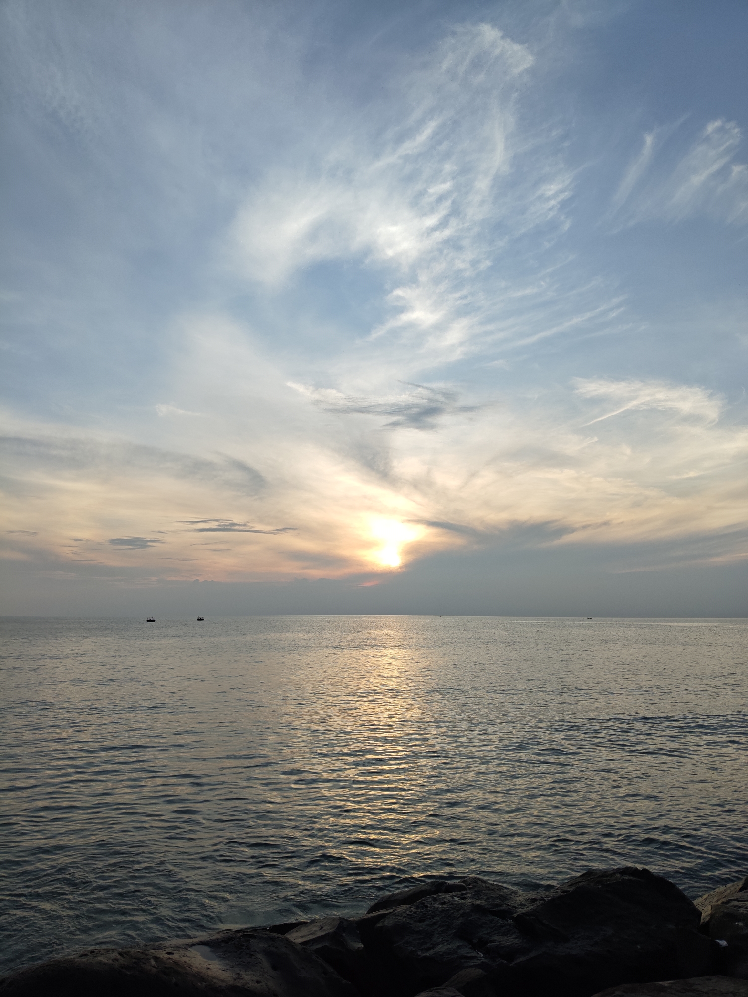 The image depicts a serene seascape with a sunset over the ocean. The sky is painted with a gradient of soft blues and oranges, with wispy clouds stretching across the horizon. The sun, a bright orb partially obscured by clouds, casts a shimmering reflection on the calm waters. In the distance, two small boats are visible, adding a sense of scale to the vastness of the sea. The foreground is occupied by dark, textured rocks, suggesting a coastline or a pier. The overall atmosphere is one of tranquility and natural beauty. This scene is likely Lhokseumawe Beach in Lhokseumawe, Indonesia, a popular spot known for its scenic sunsets.