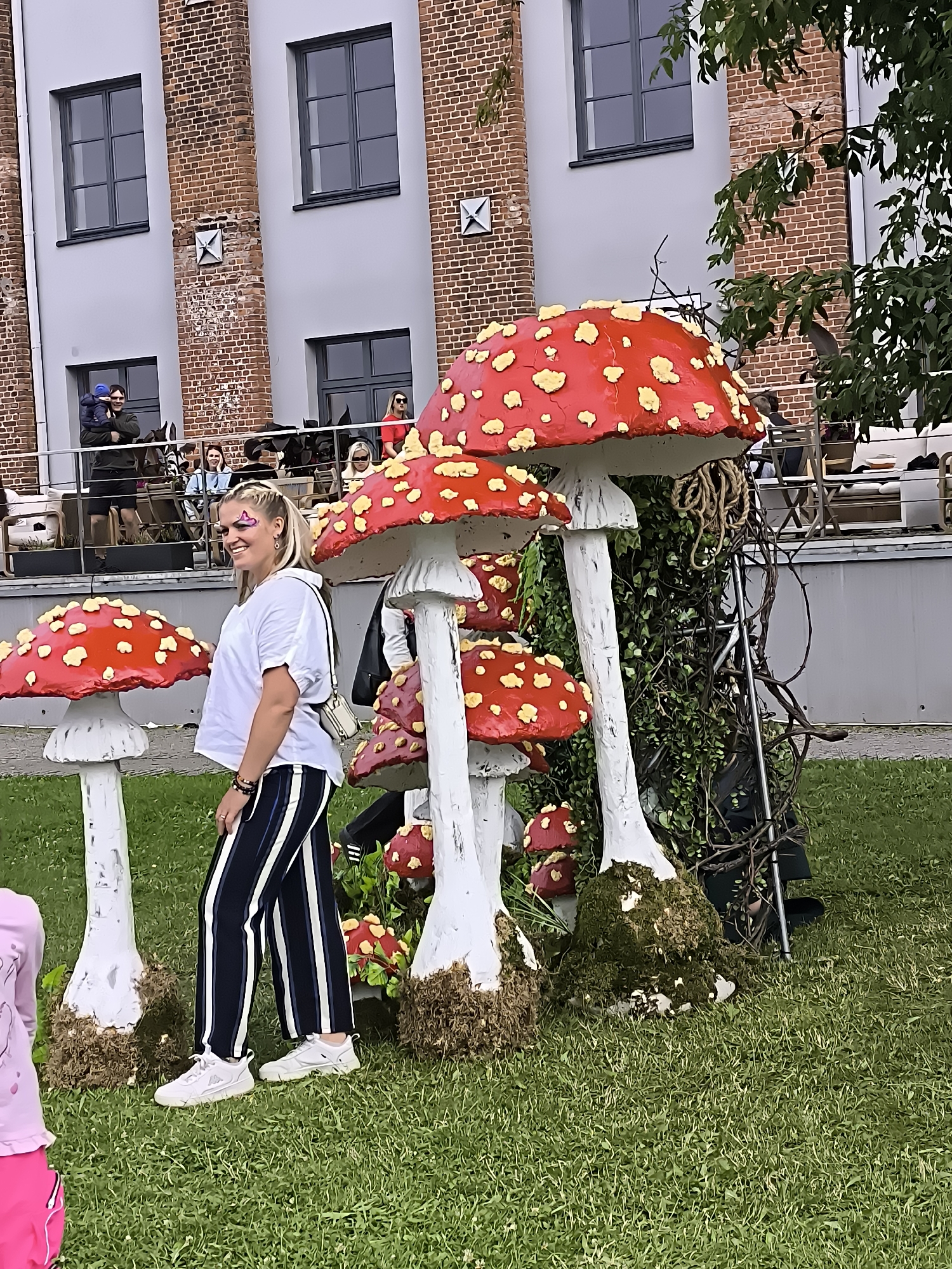 The image depicts a lively outdoor scene with a woman in the foreground, smiling and posing. She is wearing a white t-shirt, black and white striped pants, and white sneakers. Her hair is styled in braids, and she has what appears to be face paint on her cheek. She is standing among a cluster of large, decorative mushrooms with red caps and white stems, resembling fly agaric. The mushrooms are placed on a grassy area, and some are adorned with moss.
In the background, a brick building with multiple windows is visible, suggesting an urban or semi-urban environment. Several people are gathered on a balcony or elevated walkway in front of the building, some holding children. Trees with green foliage are also present, adding to the natural elements of the scene.
The overall atmosphere is festive and casual, with the woman in the foreground being the clear focal point. The decorative mushrooms suggest an event or a themed attraction.
This scene is located at the Klaipėda Old Steam Power Plant Cultural Centre in Klaipėda, Lithuania, which is known for its industrial heritage and its transformation into a vibrant cultural hub hosting various events and installations. The architectural style of the building and the presence of large-scale decorative elements are characteristic of the exhibitions and festivities often held at this location.