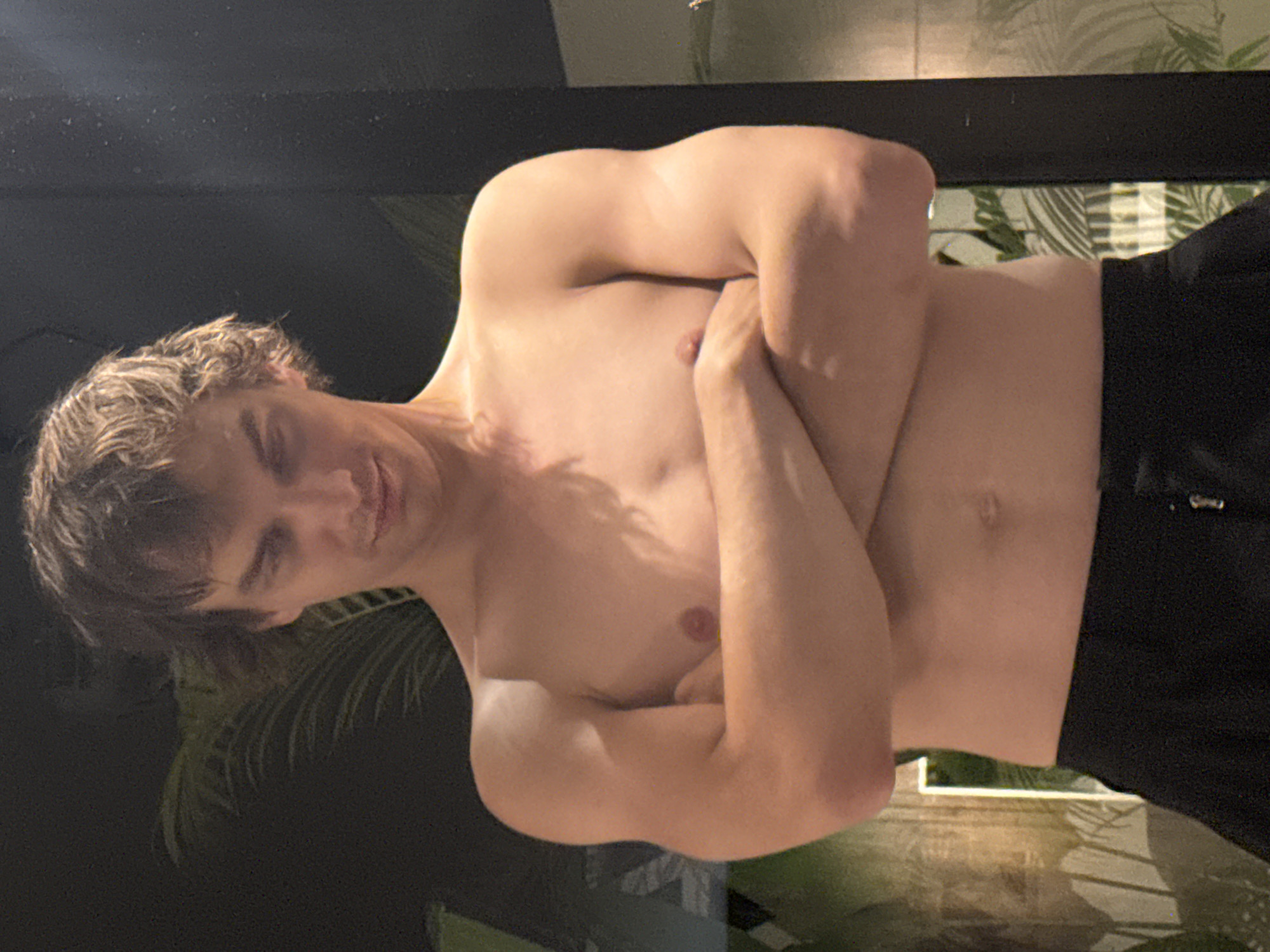 The image displays a young, light-skinned man captured from the chest up, with his head gently tilted to his left. He has short, wavy, light brown hair. He is shirtless, revealing a lean, muscular build, and is wearing dark shorts that are visible at the bottom of the frame. His arms are crossed over his chest, with his right arm positioned over his left, and his hands lightly grip his upper arms. A subtle smile or smirk plays on his lips, and his eyes are partially closed, conveying a relaxed and possibly playful expression.
The background suggests a tropical or semi-tropical environment, consistent with Kuta Selatan, Indonesia. Behind the man, a dark horizontal surface, possibly a beam or partition, is visible. Through a gap in the background, a pattern of green and white tropical leaves, resembling palm fronds, is discernible, hinting at lush foliage or a themed interior. The lower left area of the image shows a dark, reflective surface, possibly glass, wet ground, or a pool edge, with faint green reflections. The lighting is subdued, creating gentle shadows and highlights on his skin. No text is visible in the image. The scene evokes a calm, private atmosphere.