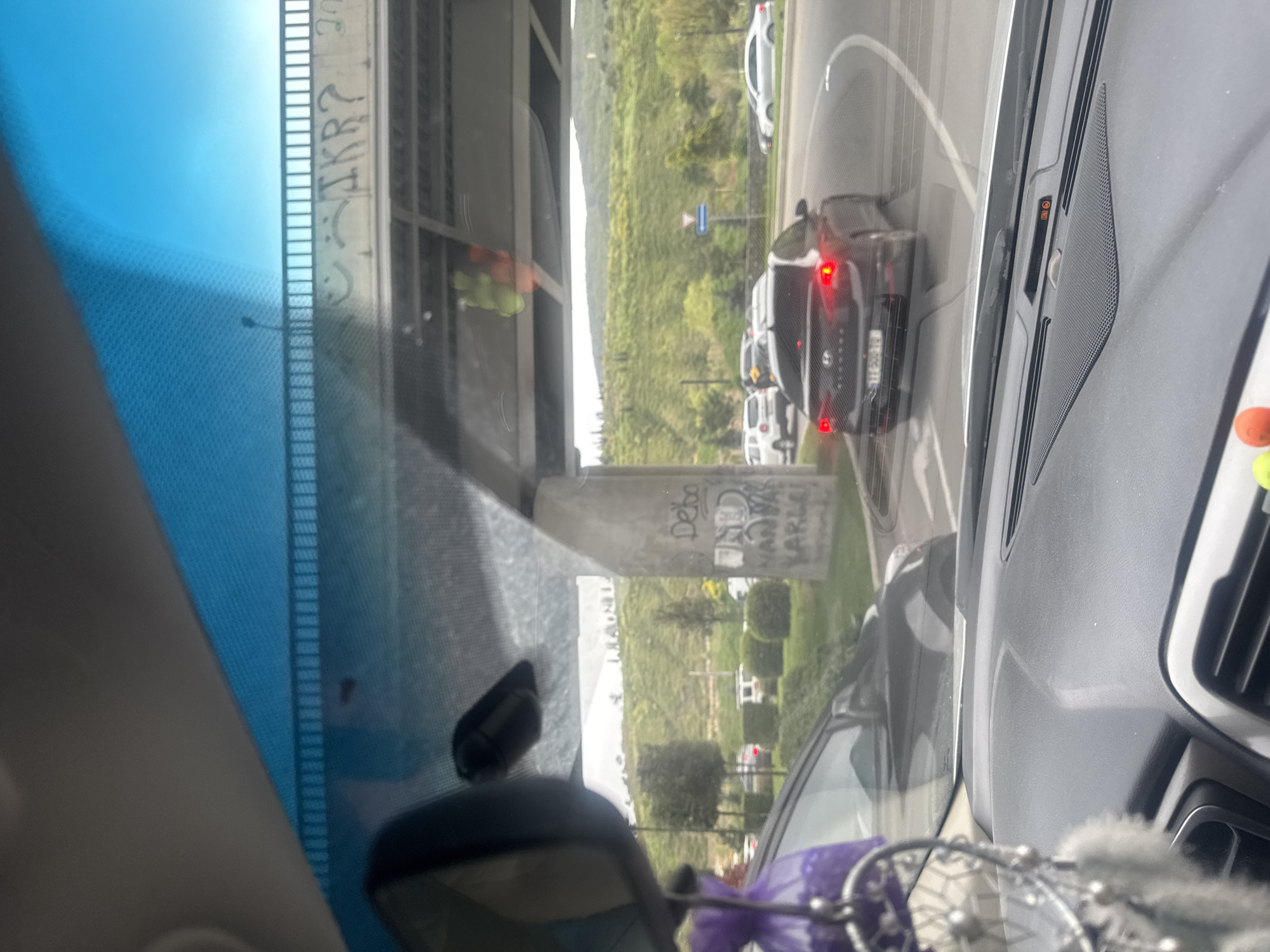 The image captures an outdoor scene from the interior of a car, likely on a road in Tbilisi, Georgia, on a clear, bright day. The perspective is through the front windshield, looking out at traffic and infrastructure.
In the foreground, elements of the car's interior are visible, including parts of the dashboard, a speaker grille on the right, and an orange warning light (possibly an airbag indicator). A decorative item, resembling a dreamcatcher, hangs from the rearview mirror in the lower right corner. The windshield shows reflections and glare, particularly in the upper left.
Ahead on the road, traffic appears to be stopped or moving slowly, indicated by the illuminated red brake lights of a dark-colored SUV or crossover directly in front. Other white cars are visible further down the road. A blue rectangular road sign, possibly a yield sign, stands on the roadside. The road seems to curve towards the right.
A significant feature is a concrete overpass or bridge structure that spans the road. Graffiti marks its surfaces. On an upper beam, the letters "JKR?" are visible. On a lower concrete support pillar, the word "DeKo" is clearly spray-painted, with additional, less distinct text below it, including "OUD" or "OND" and "WARLI" or "WARLLI".
Beyond the road and concrete structures, a verdant, hilly landscape extends into the distance, suggesting natural terrain or mountains. The sky is a clear blue, indicating good weather conditions. The scene portrays a blend of urban infrastructure and natural environment typical of areas like Tbilisi.
