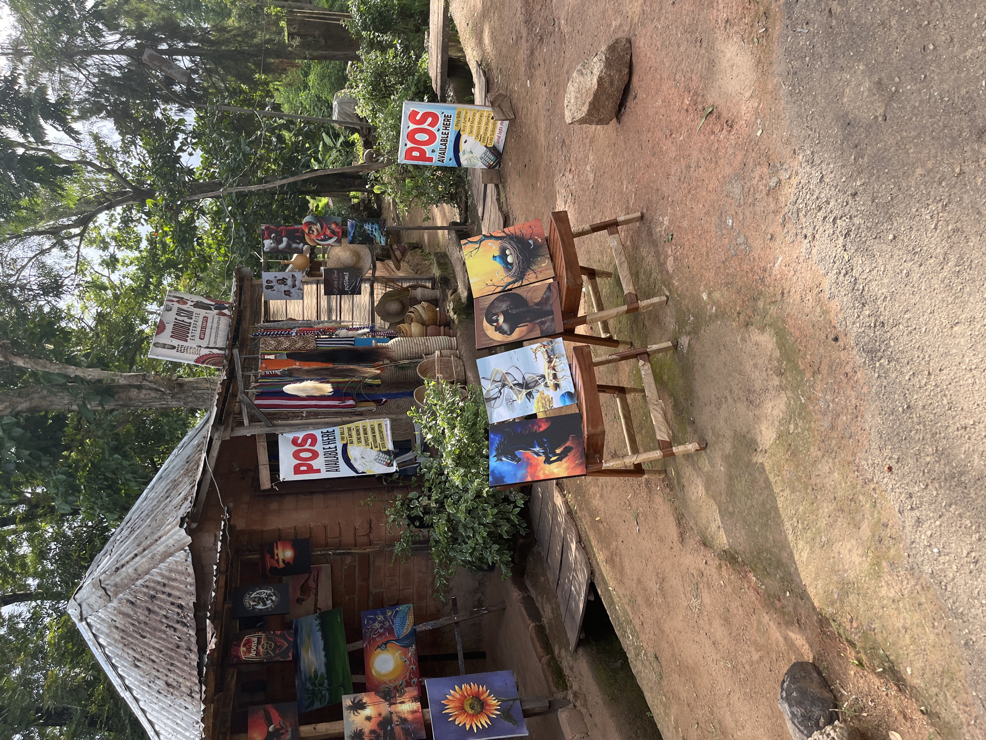 The image depicts an outdoor art stall in Jos, Nigeria, exhibiting paintings of various subjects including wildlife, sunsets, and a sunflower. Several artworks are displayed on easels and propped against the stall. A prominent sign reads "POS AVAILABLE HERE," listing services such as paying bills, buying airtime, sending, and withdrawing money. The stall itself is constructed from brick and has a corrugated metal roof. The surrounding environment is characterized by lush green trees and foliage, with a dirt path in the foreground. The scene conveys a typical market or roadside vendor setup in a rural or semi-urban area.