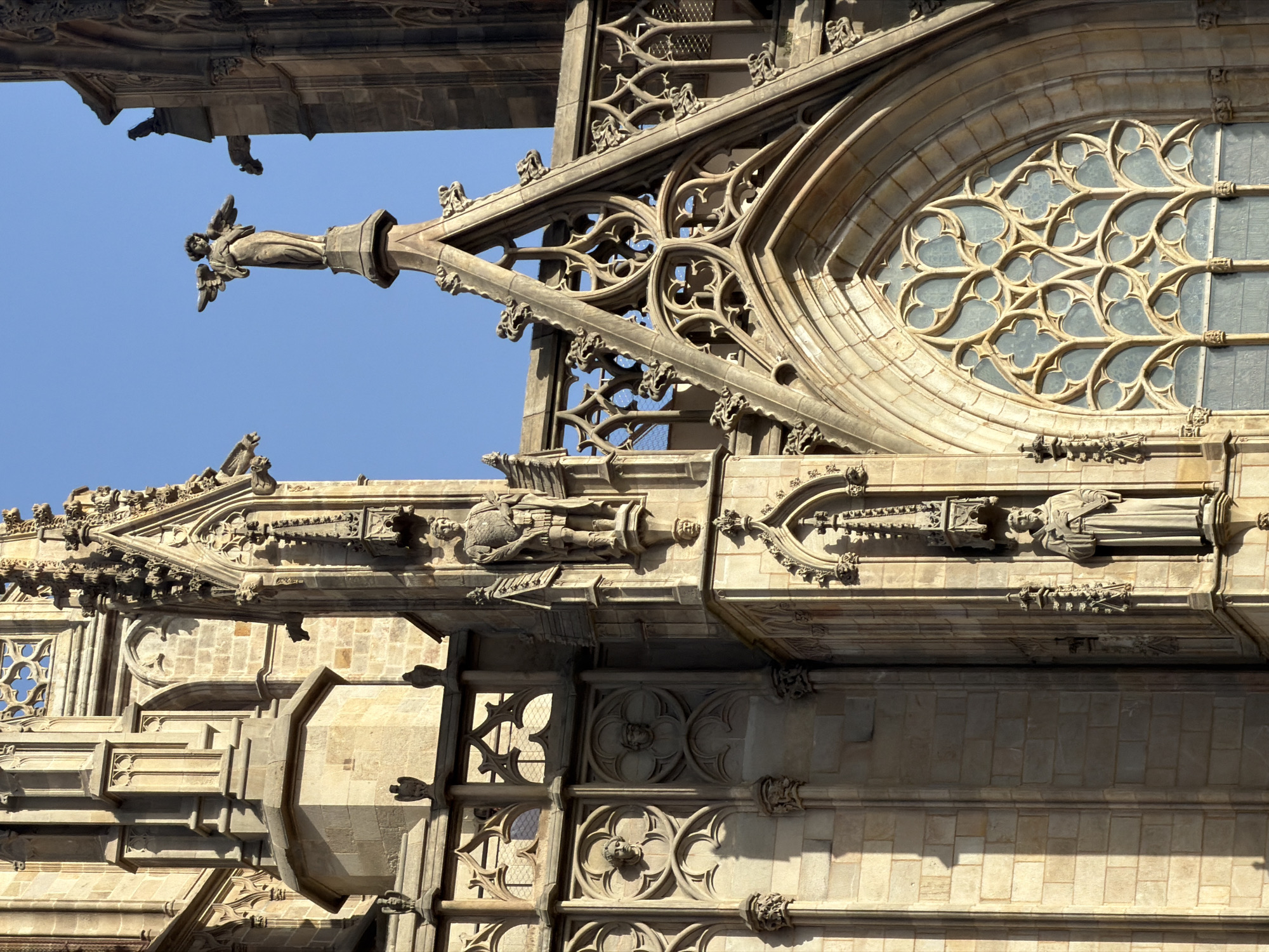 The image shows a close-up, detailed view of the Gothic facade of the Barcelona Cathedral, located in the Gothic Quarter of Barcelona, Spain. The architectural style is characterized by intricate stonework, pointed arches, and decorative tracery.
Dominating the upper portion of the image is a statue of an angel with outstretched wings, standing atop a spire. Below this, and to the left, are niches containing two stone statues of robed figures, one appearing to be a soldier or dignitary in armor, and the other a monk holding a book. The stonework of the cathedral is a warm, sandy color, illuminated by sunlight, creating distinct shadows.
Various Gothic architectural elements are visible, including ornate window tracery, gargoyles, pinnacles, and decorative moldings. The background is a clear, bright blue sky. The overall impression is one of grandeur and historical significance, highlighting the detailed craftsmanship of medieval architecture.
