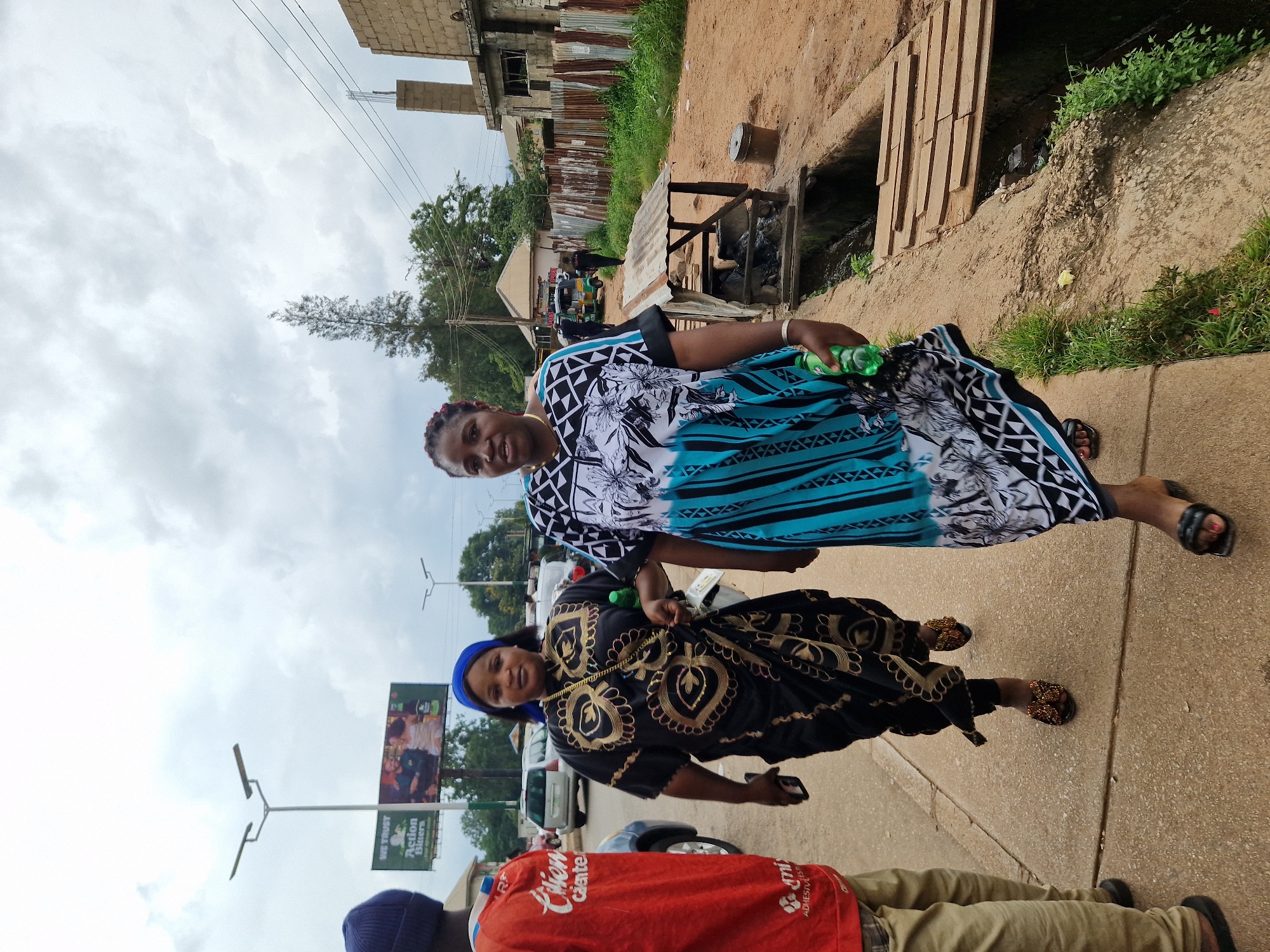 In Jos, Nigeria, two women are captured in a full-body, eye-level shot as they walk side-by-side on a paved sidewalk next to a dirt road. The woman on the left wears a black and gold patterned dress and a blue headscarf. The woman on the right is dressed in a blue and white patterned kaftan-style dress. Both women are looking towards the camera and appear to be engaged in conversation. The woman on the right is holding a green plastic bottle in her right hand.
The background features a typical urban street scene in Jos. Several cars are parked or moving on the road, including a silver sedan and a Bajaj auto-rickshaw. A man in a red shirt is visible in the foreground on the left, partially obscuring the view of the woman in the black dress. In the distance, buildings under construction, a billboard, and utility poles are visible. The area also includes a partially covered drainage ditch with a wooden walkway crossing it, and a rustic shelter with a corrugated metal roof. The sky is overcast, with scattered clouds indicating a typical weather pattern for the region. The overall scene conveys a sense of everyday life in a Nigerian city.