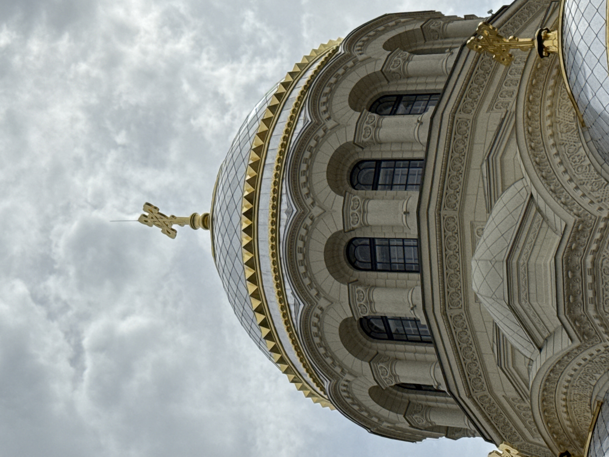 The image shows a close-up, low-angle shot of the dome of the St. Nicholas Naval Cathedral in Saint Petersburg, Russia. The dome is ornate, with gold accents and a golden cross at its apex. The cathedral's facade is light-colored stone, featuring arched windows and intricate carvings. The sky above is overcast with dramatic, textured clouds. No people are visible in the frame. The image focuses on the architectural details of the cathedral against the backdrop of a cloudy sky.