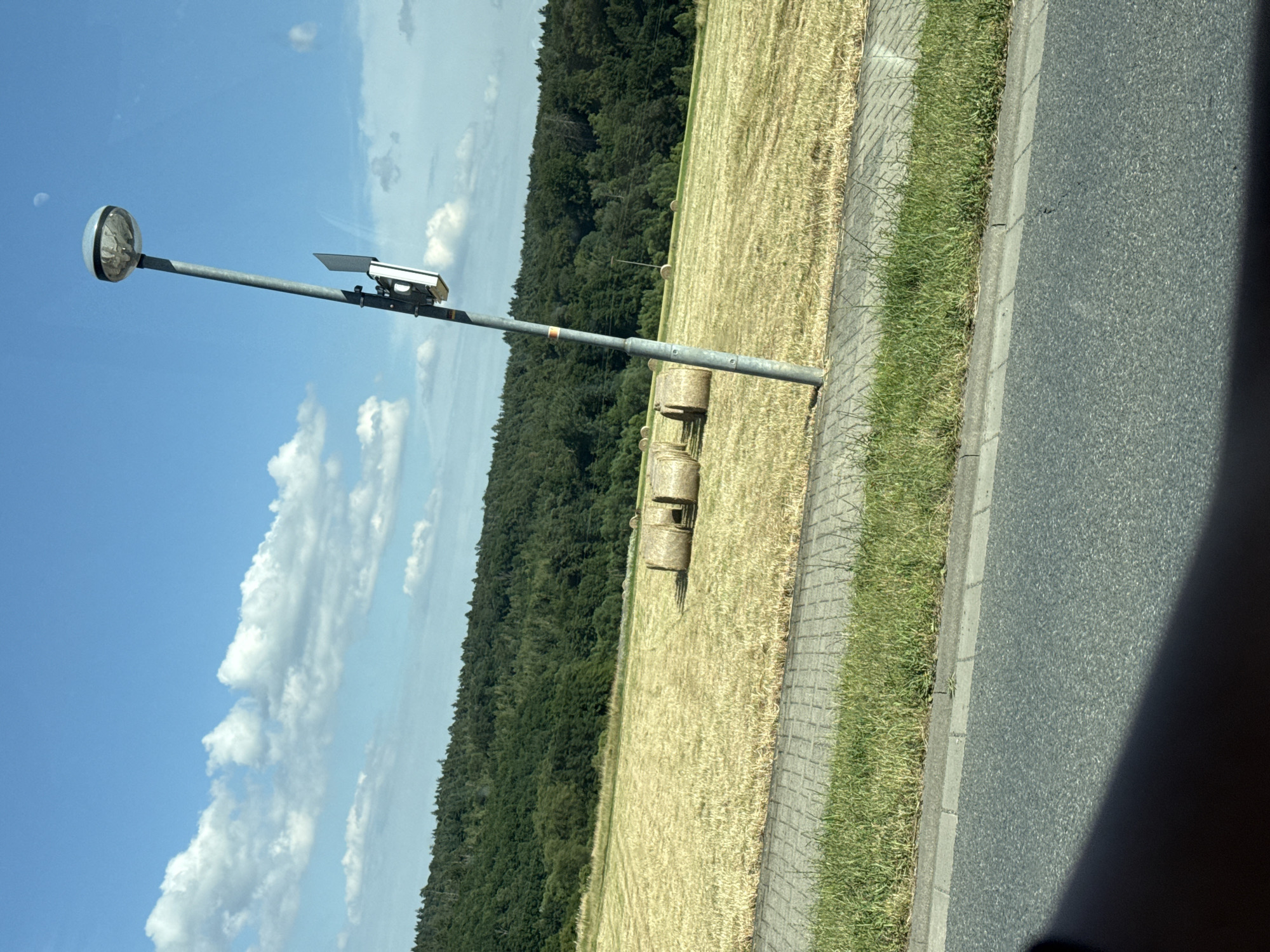 This media file shows a street-side scene in Volpertshausen, Germany. The primary objects are a metal pole with a circular light fixture at the top and a device mounted halfway down, likely a traffic camera or sensor. In the background, a field of harvested wheat, indicated by straw bales, stretches towards a dense, green forest. The scene is bordered by a paved sidewalk and an asphalt road. The sky is bright blue with scattered white clouds, suggesting a clear, sunny day, possibly late morning or early afternoon. There is no visible text in the image. The overall impression is one of a rural or semi-rural roadside setting during harvest season.