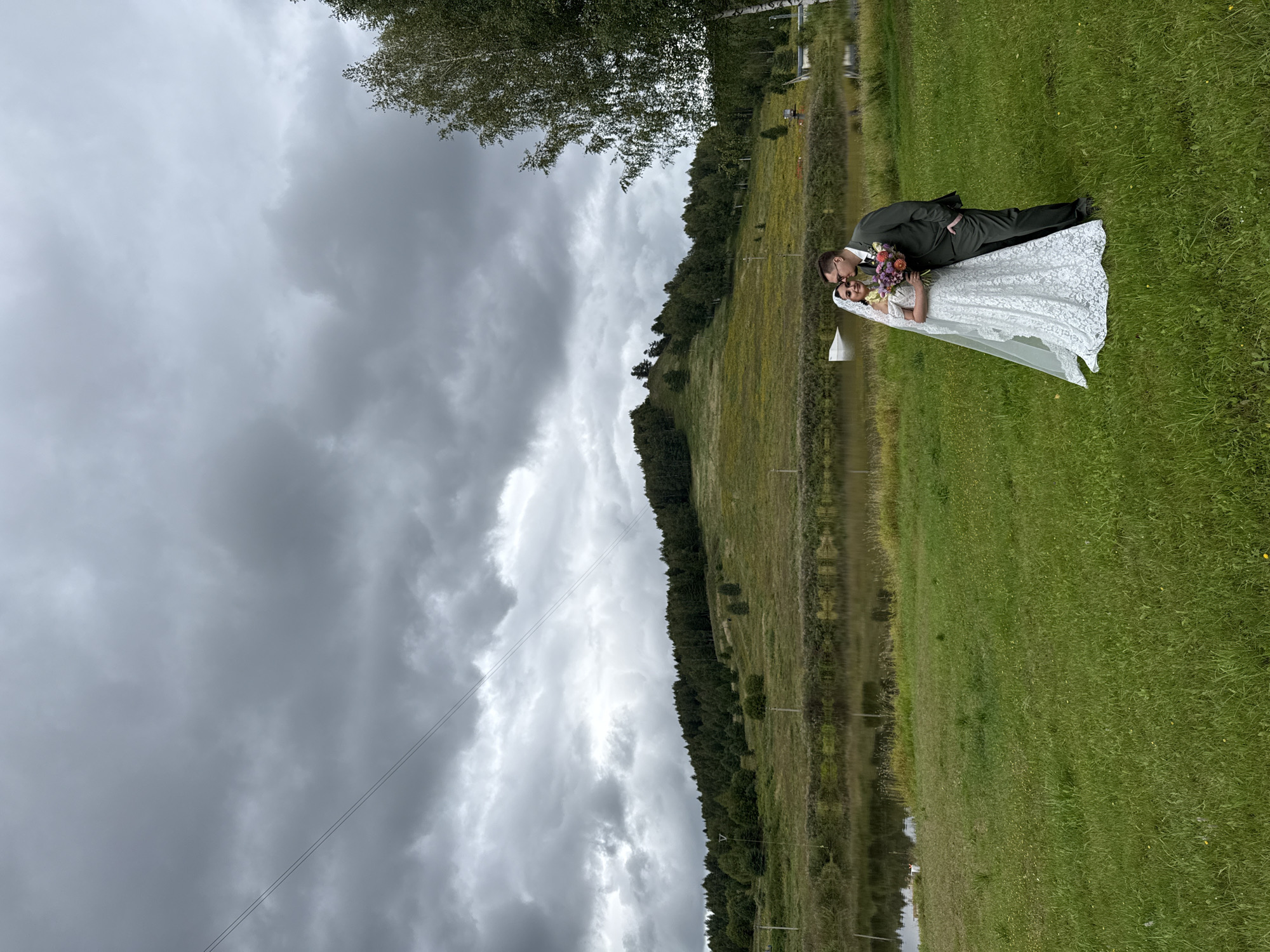 A wedding couple, a bride in a white lace dress and veil, and a groom in a dark suit, stand on a grassy field. The groom is kissing the bride's cheek. They are positioned in the foreground to the right of center. Behind them is a lake with reflections of the overcast sky and trees. In the background, a tree-covered hill rises steeply, with some patches of yellow flowers and bare earth visible on its slopes. Power lines are visible in the sky above the hill. The overall scene depicts a wedding ceremony or photoshoot in a natural, somewhat somber outdoor setting. The location is identified as Pechmen', Russia.