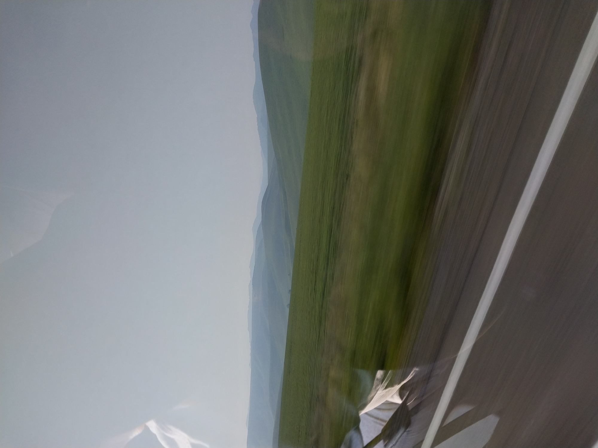 The image depicts a hazy, rolling landscape of green hills under a pale sky. The foreground shows a blurred view of a road with a white line, suggesting motion. In the reflection on the left side of the image, a person's head and shoulder can be faintly seen, wearing white clothing. The scene appears to be captured from a moving vehicle. The location is identified as Kamyshenka, Russia.