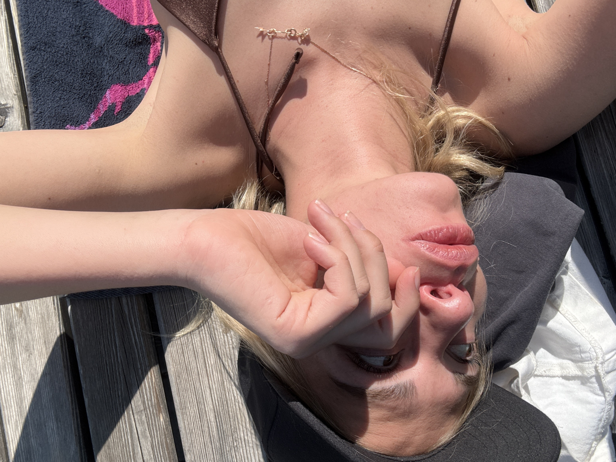 This image features a young individual, likely a woman with blonde hair, reclining on a wooden surface under bright, direct sunlight. The perspective is unusual, with the person lying on their back and looking upwards towards the camera, making their face appear upside down relative to their body.
The subject is wearing a brown bikini top and a delicate gold-colored necklace with small charms. A dark grey or black cap is partially visible on their head. One hand is raised, gently covering part of their face, possibly to shield from the sun or in a moment of relaxation. Their lips are slightly parted, and their eyes are open, gazing upwards.
The setting appears to be an outdoor wooden deck or pier, indicated by the distinct parallel planks. Strong sunlight casts sharp shadows across the wooden surface, suggesting a clear, sunny day, likely midday or early afternoon. To the left, a dark-colored towel with a striking pink, abstract pattern is visible, draped over the wood and partially under the person's arm. Other fabrics, including dark grey and white material, are seen around their head and body, suggesting they are comfortably settled.
The scene conveys a sense of leisure and warmth. While the provided context indicates the location as Krasnoyarsk, Russia, there are no specific visual cues within the image itself to confirm this geographical detail. No discernible text is present in the image.