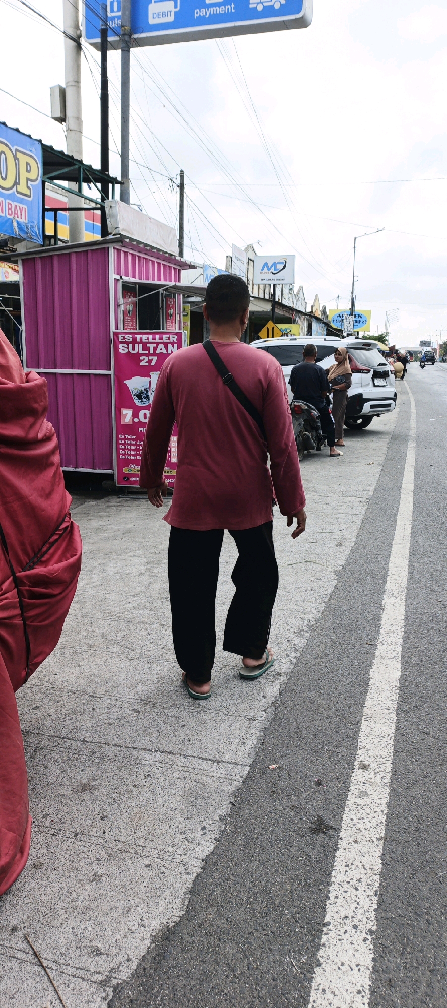 A man with short dark hair walks away from the camera on a concrete sidewalk. He is wearing a long-sleeved maroon shirt and black pants, and light-colored flip-flops. A black strap is slung across his chest. To his left is a pink stall with the text "ES TELLER SULTAN 27" and pricing information. To the right of the sidewalk is a street with white lane markings. Further down the street, a white SUV is parked, and two people are standing near it, one on a motorcycle. In the background, there are various signs and a telephone pole. The sky is overcast. The scene appears to be a street-side commercial area in Kasiyan, Indonesia.
