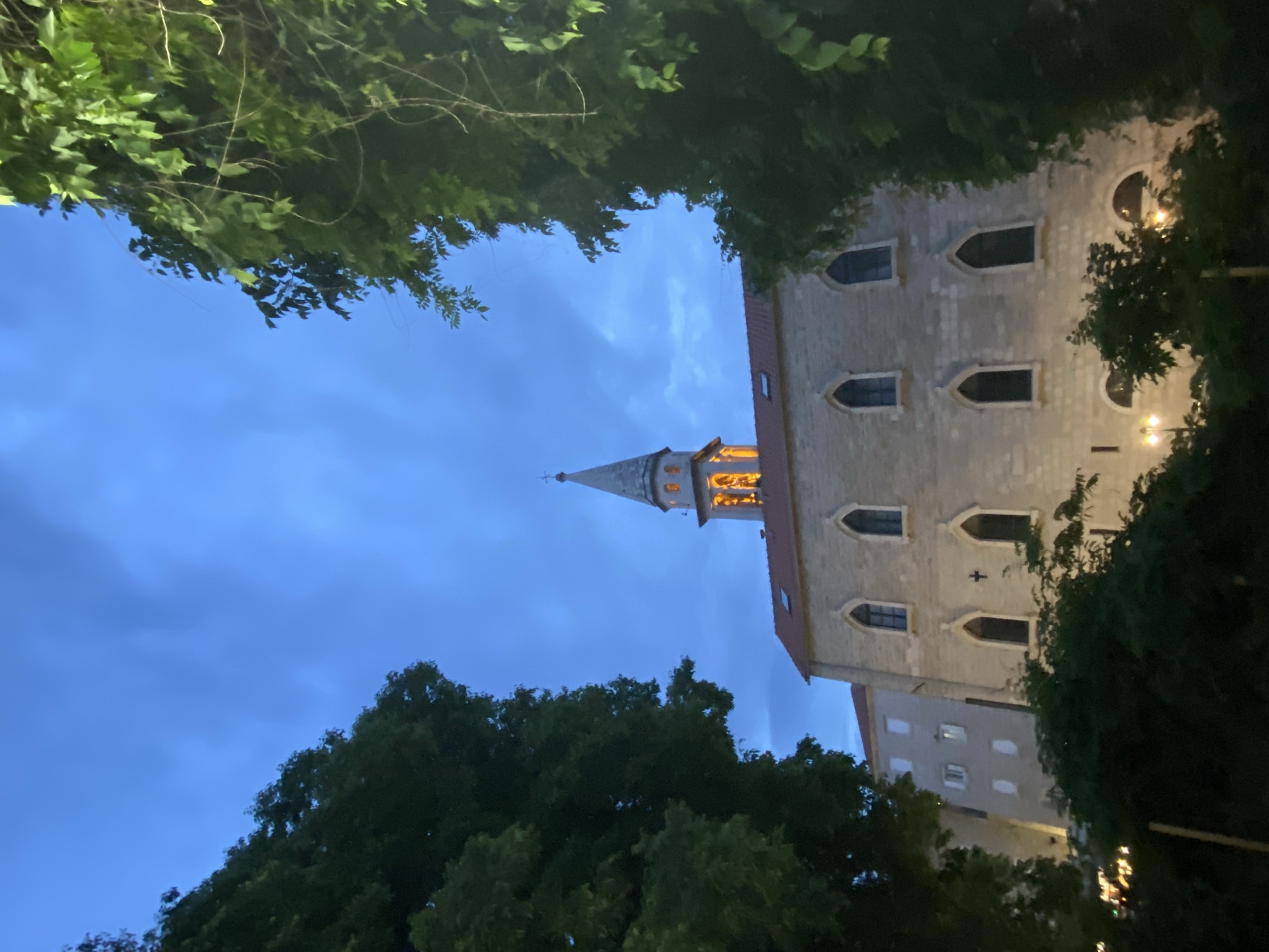 The image captures a view in Budva, Montenegro, at dusk or night. A stone building with a tall, pointed spire is the focal point, framed by tree foliage in the foreground. The spire is illuminated, likely by interior or external lights. The building has arched windows and is of a light stone color. Other buildings are visible adjacent to the primary structure. The sky is a deep blue with cloud cover. The trees in the foreground are dark green, creating a frame for the architectural details.