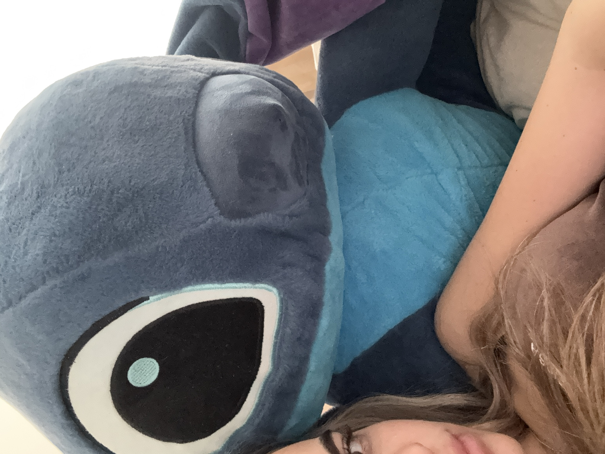 The image is a close-up, angled shot featuring a person and a large plush toy, likely captured indoors. The dominant subject is a blue plush toy resembling Stitch from Disney's Lilo & Stitch, occupying the left and upper portions of the frame. Its large, embroidered eye, with a black pupil, white sclera, and a light blue highlight, is prominent. The toy has a soft, fuzzy texture, and other parts of its body, including a purple ear and lighter blue sections, are visible in the background.
In the bottom right quadrant, part of a person's face is visible, including their nose, mouth, and chin, suggesting they are lying down. Light brown hair with blonde highlights frames their face and extends towards the center of the image. An arm or shoulder, showing some freckles, is also visible to the right, resting near the plush toy.
The background consists of various shades of blue and purple fabric, likely blankets, pillows, or other soft furnishings, indicating a comfortable setting such as a bed or couch. A light-colored surface, possibly a wall or bedding, can be seen in the upper left.
The scene conveys a sense of comfort and relaxation, with the person appearing to be resting or cuddling with the large plush toy. There is no visible text. The time of day and weather are not discernible from the image. The location context is Sofia, Bulgaria.