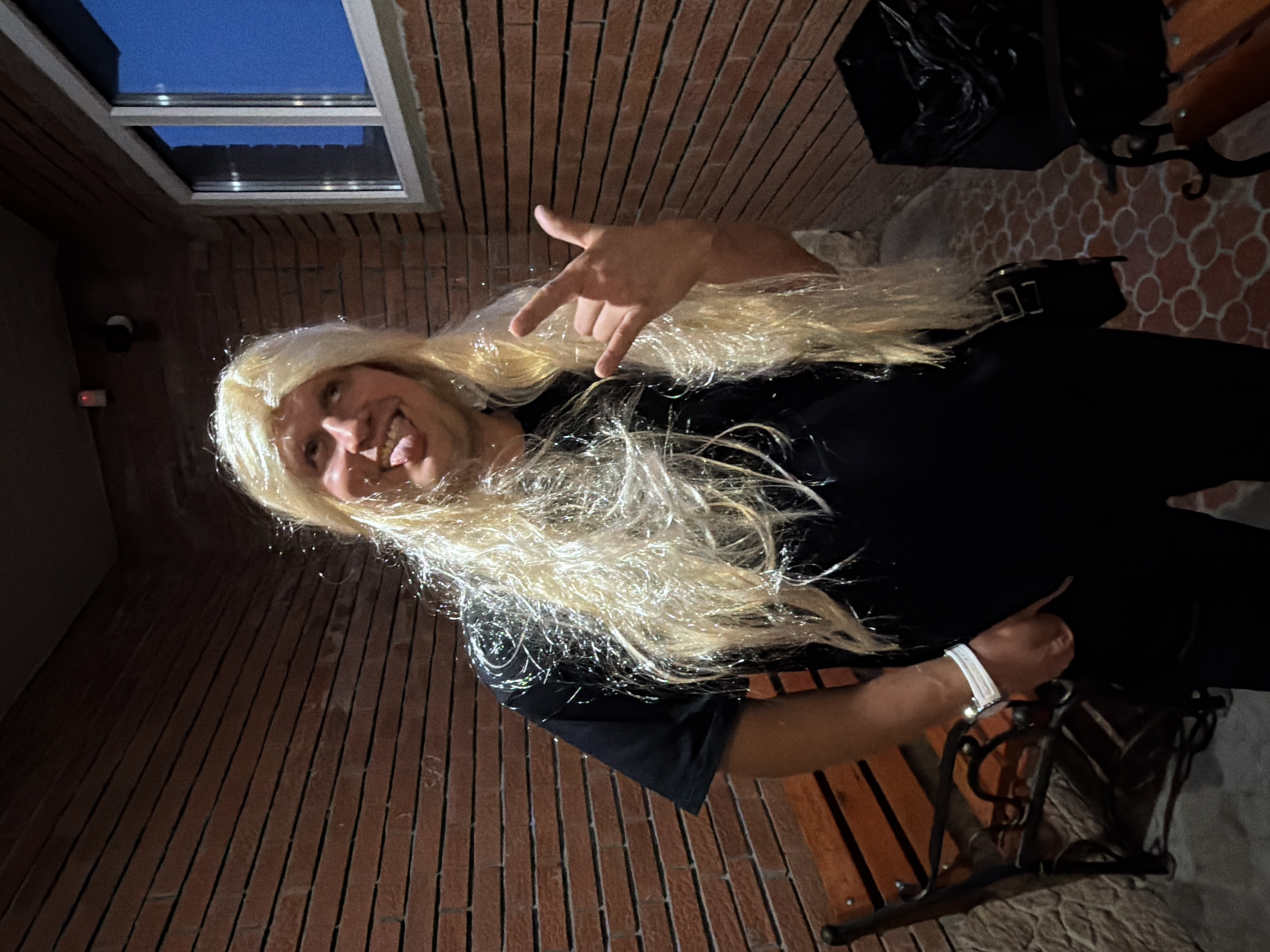 In Chaikovsky, Russia, a person wearing a blonde wig with shimmering strands and a black t-shirt stands in front of a brick wall. They are sticking out their tongue and making a rock and roll hand gesture. To the left, a wooden bench with a black metal frame is visible, and to the right, a black trash can with a plastic bag liner. The ground is paved with circular brick patterns. A window with a blue sky reflection is also present on the brick wall.
