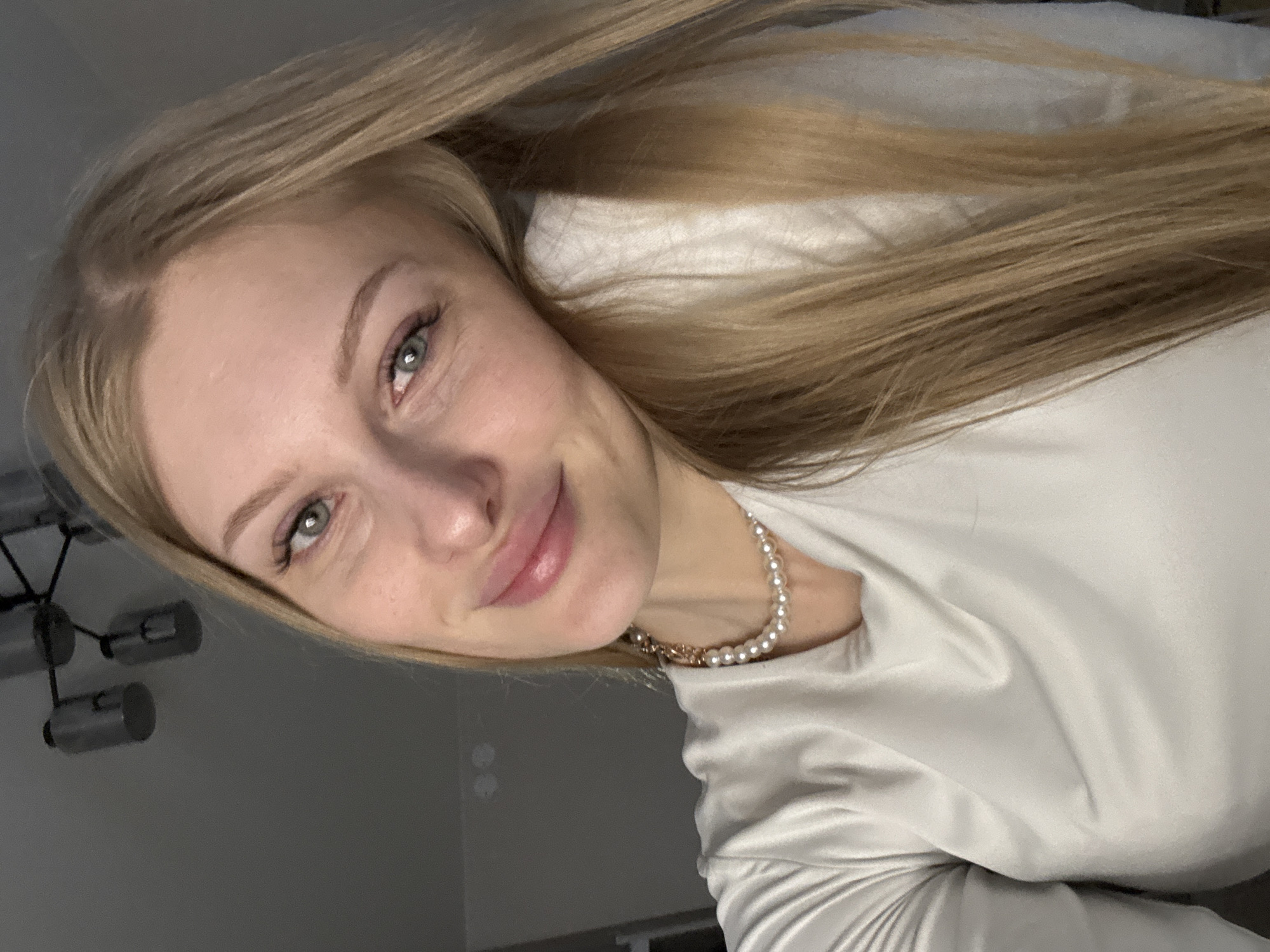 A portrait-style image, rotated counter-clockwise, depicts a young woman with long, straight, light blonde hair. Her head is tilted slightly, and she looks directly at the camera with light greenish-blue eyes, displaying a soft, closed-mouth smile. She has fair skin and light, well-defined eyebrows.
She is wearing a light-colored, possibly off-white or light grey, top made of a satin-like fabric. A pearl necklace with a visible gold chain segment is draped around her neck.
The background indicates an indoor setting. To the left side of the frame, a modern light fixture with multiple cylindrical elements is visible against a dark grey wall. Behind her head and flowing hair, a light-colored, soft surface, likely a pillow or bedding, is present. The overall lighting is soft and consistent, typical of indoor illumination.
The woman's expression conveys a sense of calm and pleasantness, suggesting she is relaxed while posing for the photo. There is no visible text in the image. This scene takes place in an indoor setting, consistent with Perm, Russia.