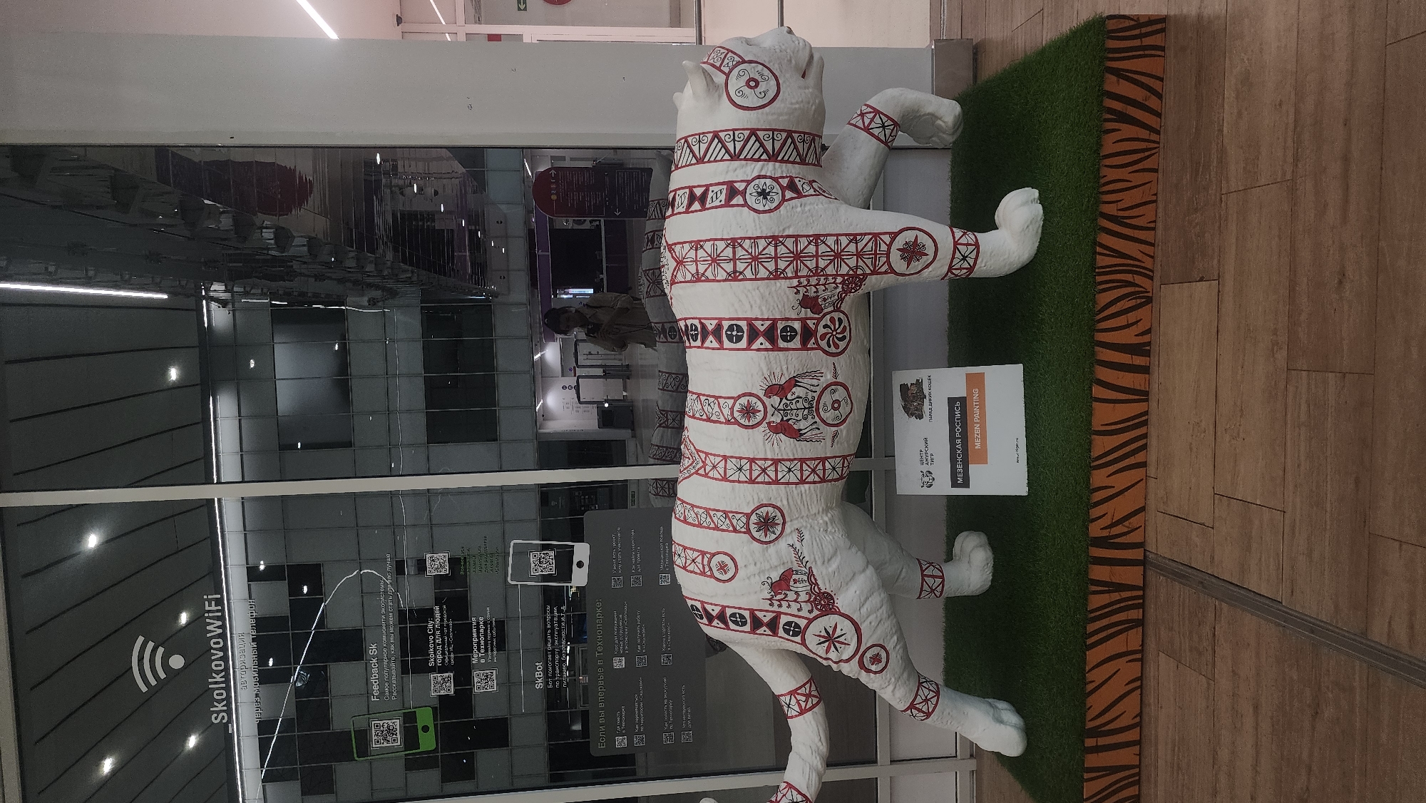 The image shows a white tiger sculpture decorated with red and black traditional Russian patterns, specifically Mezen painting. The sculpture is placed on a green artificial grass mat, which is on a platform with a tiger-striped pattern. Behind the sculpture, there is a large glass window reflecting the interior of a modern building, possibly a shopping mall or an exhibition space. The reflection shows a woman taking a photo and part of the building's architecture with lights. Signage in Russian is visible on the glass, including "Skolkovo WiFi" and information about Skolkovo City and events. Below the sculpture, a sign in Russian and English reads "МЕЗЕНСКАЯ РОСПИСЬ" and "MEZEN PAINTING" along with logos of "ЦЕНТР АМУРСКИЙ ТИГР" and "ПАРАД ДАЙКОВЫХ КОШЕК". The overall scene suggests an art installation or an exhibition related to Russian culture and wildlife conservation, possibly located within the Skolkovo Innovation Center in Moscow, Russia.