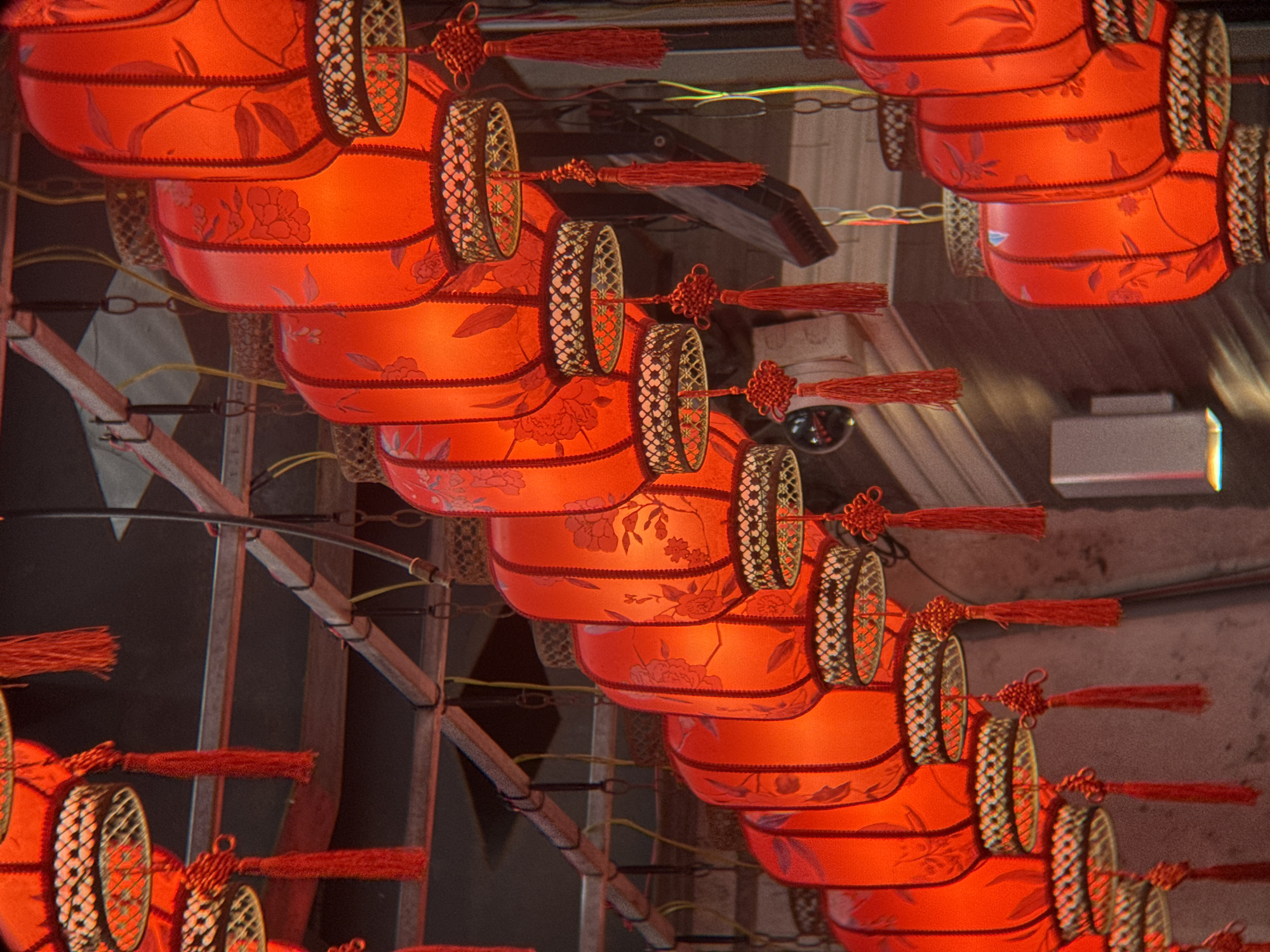 This media file displays a close-up view of several traditional red Chinese lanterns, possibly hung for decoration or a festive occasion. The lanterns are cylindrical with intricate floral patterns etched onto their surfaces. They are adorned with decorative red tassels and detailed golden or brass-colored rings with a patterned mesh. The lanterns are arranged in rows, hanging from above, creating a visually dense composition. The lighting suggests an indoor or evening setting, with the red glow of the lanterns being the primary light source. The background is dark and out of focus, with hints of metal structures, possibly part of the hanging mechanism or the surrounding architecture, and a faint glimpse of what appears to be a surveillance camera. The scene evokes a sense of cultural celebration or traditional ambiance. No text is visible in the image. The location context is Nha Trang, Vietnam.