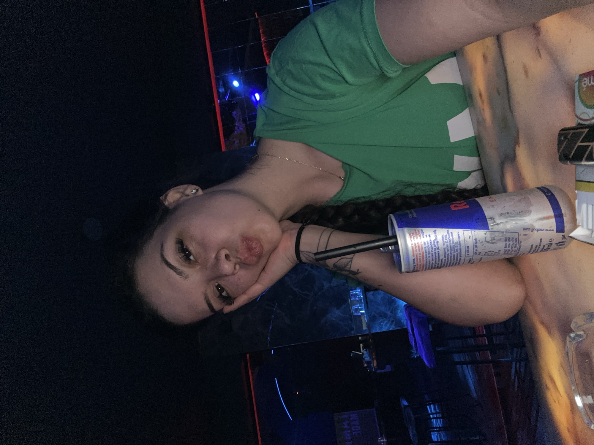 A young woman with dark hair braided down her back is taking a selfie in a dimly lit establishment. She is wearing a bright green t-shirt with a white graphic design. Her left hand is propping up her face, and she is puckering her lips. In front of her on a wooden surface is a can of Red Bull and an ashtray. The background appears to be a bar or club setting with blue and red neon lights and dark furniture. The location is identified as Sliven, Bulgaria.