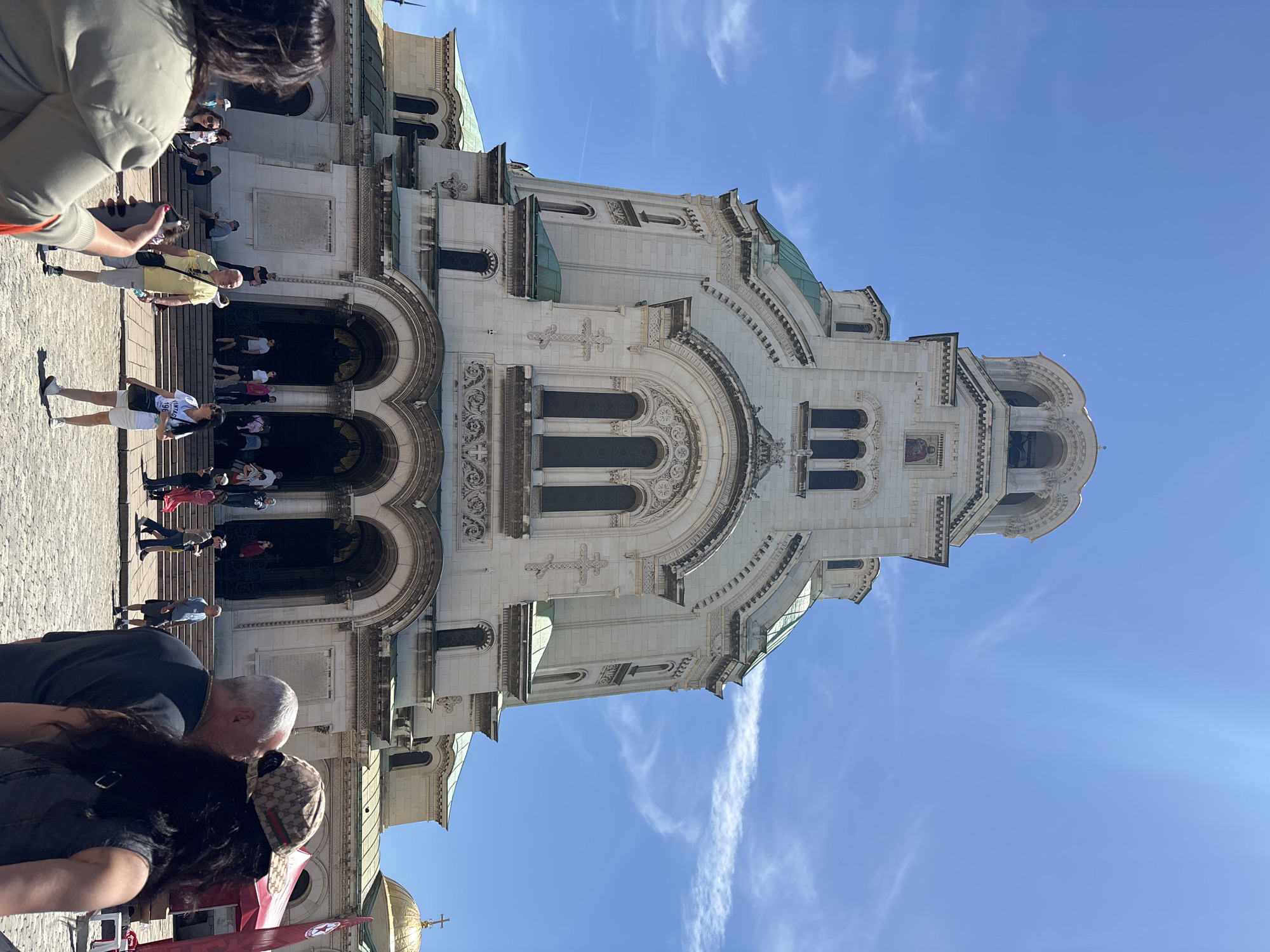 The image captures the Alexander Nevsky Cathedral in Sofia, Bulgaria, seen from a low angle. The cathedral is made of white stone and features intricate architectural details, including arched windows, decorative moldings, and crosses. In the foreground, a couple is kissing, with the woman wearing a patterned hat. To the left, there is a crowd of people standing on the steps leading to the cathedral entrance. The sky is a clear blue with scattered wispy clouds. A golden dome with a cross is partially visible at the bottom left corner.