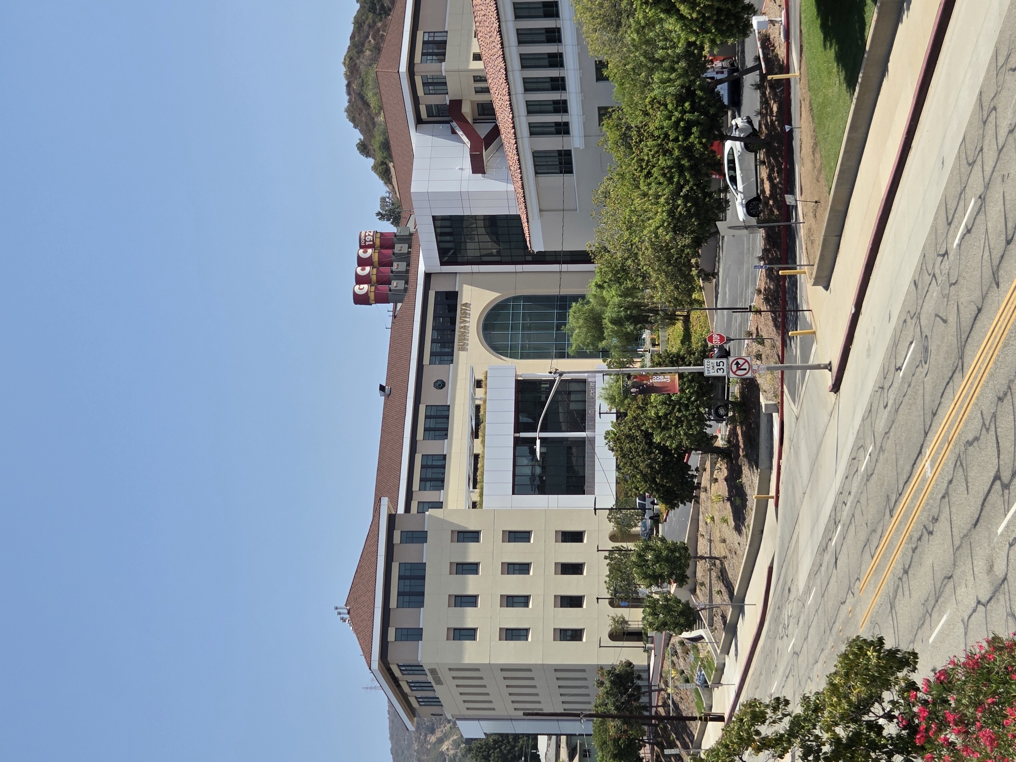 The image captures a wide-angle view of the Glendale Community College campus in Glendale, California, United States of America. The scene is dominated by a modern, multi-story building with beige and white facades and a reddish-brown tiled roof. Large windows, some tinted, are visible on all levels of the building, reflecting the bright, clear blue sky. Atop the building, prominent red cylindrical structures with white lettering that reads "GCC" and "FOUNDED 1927" are visible.
In front of the building, a paved road with clear lane markings and a red curb runs diagonally from the foreground to the midground. Several trees with green foliage are planted along the roadside and on a raised, landscaped area adjacent to the road. A white SUV is parked on the right side of the road, near a grassy area. Road signs, including a speed limit sign indicating "35" and a stop sign, are positioned near the road.
The background features a hillside covered in green vegetation, typical of the Southern California landscape. The overall impression is one of a well-maintained, institutional environment on a sunny day. No people are clearly visible in the image.