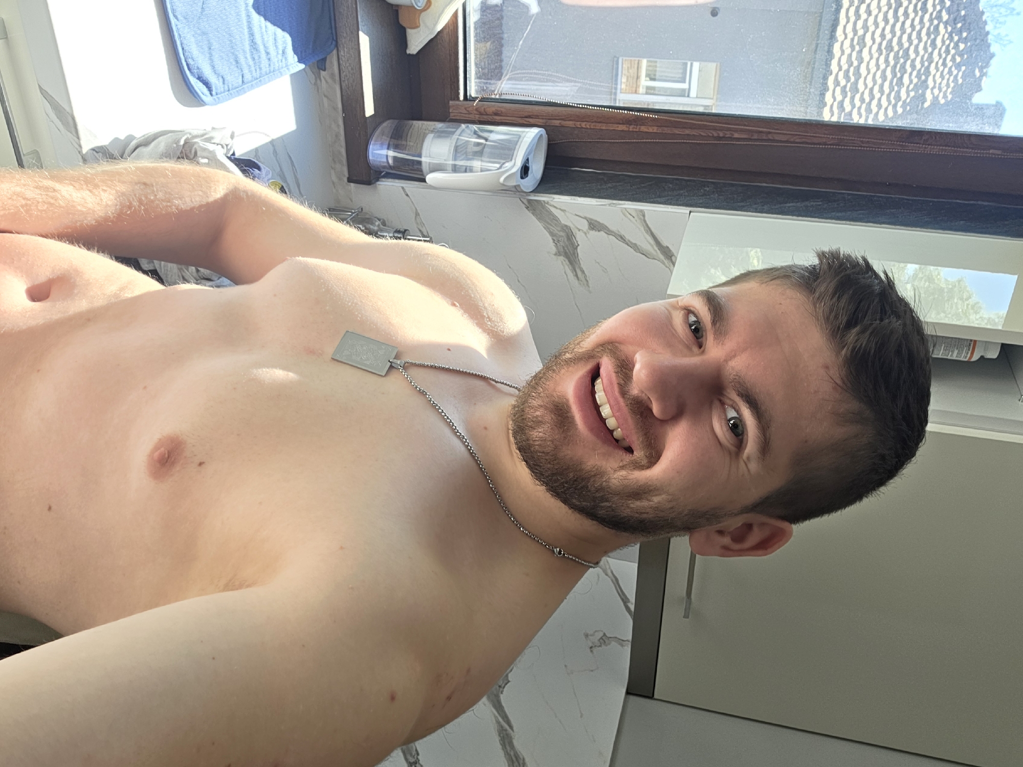 A shirtless man with a beard and short dark hair is smiling at the camera. He is wearing a silver necklace with a rectangular pendant. The background includes a window with a glimpse of a building with a roof, and a partially visible countertop with items like a water bottle. The location cannot be determined to be Yagodovo, Bulgaria, as there are no identifying features.