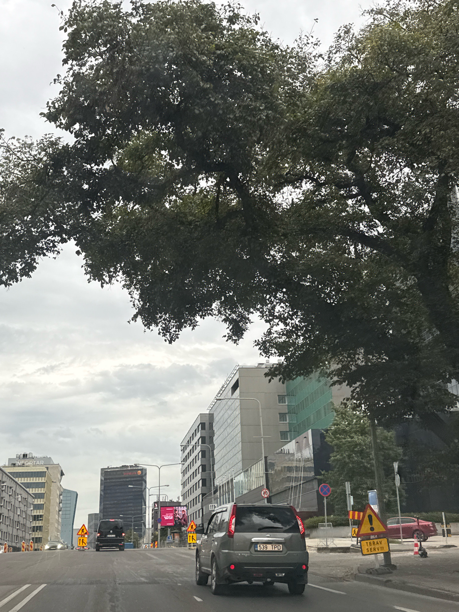 The image captures a street view in Tallinn, Estonia, likely on a cloudy day. A large tree with dense dark green foliage dominates the upper portion of the frame, partially obscuring the sky. In the foreground, a dark grey SUV is driving on a multi-lane road. The SUV has a license plate that reads "539 TPD". Further down the road, a black van is also visible. The street is lined with modern buildings of various architectural styles and heights. Some buildings feature reflective glass facades, while others are clad in lighter materials. Construction barriers and traffic signs, including warning signs with yellow triangles and pedestrian symbols, are present along the road, indicating potential roadwork or temporary diversions. A large digital billboard displaying an advertisement is visible on the right side of the road. A red sedan is parked on the right shoulder of the road, near a traffic cone and a scooter. The overall impression is of an urban environment with ongoing development or maintenance.