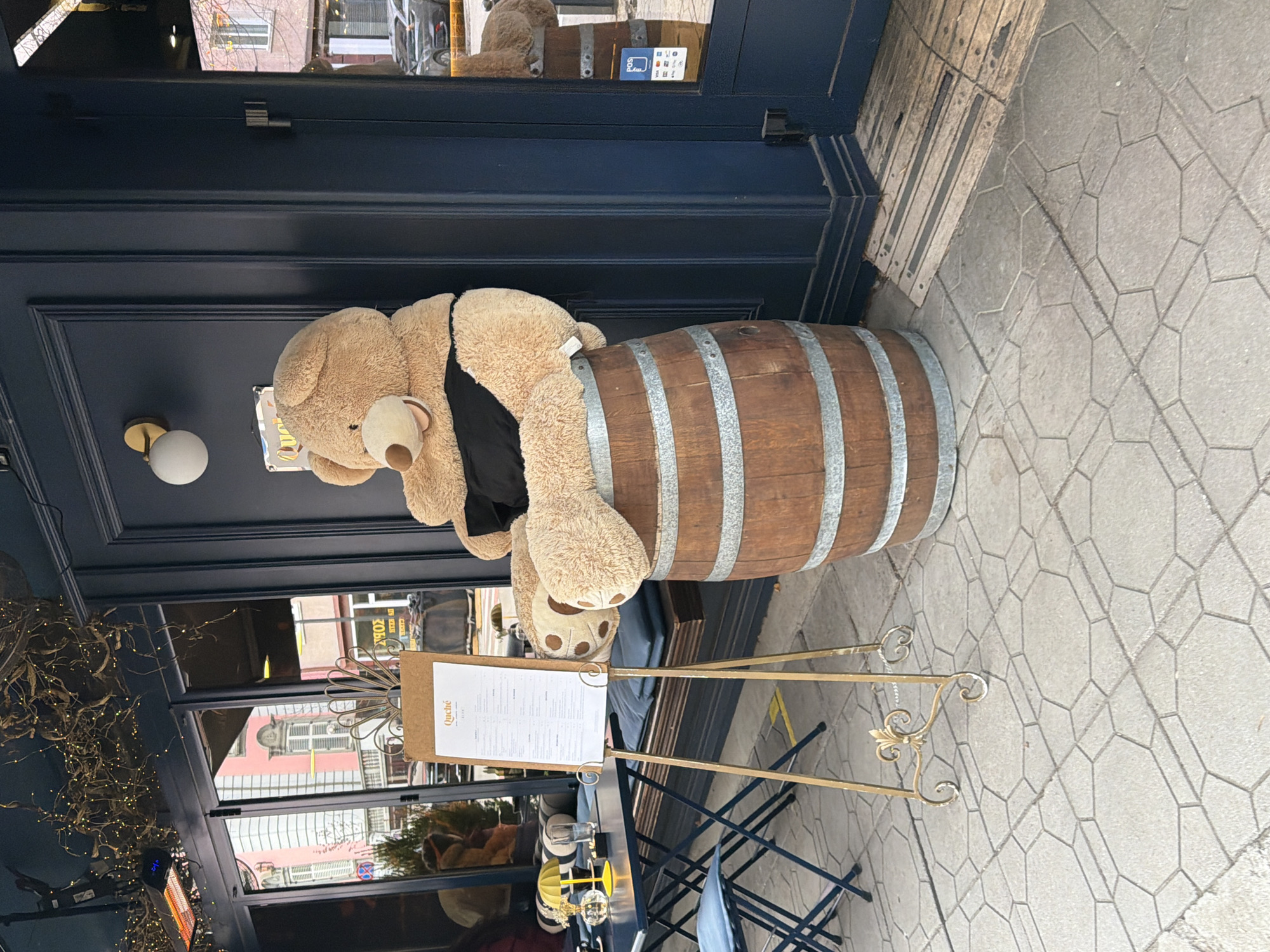 A photograph depicts a teddy bear draped over a wooden barrel outside a restaurant in Sofia, Bulgaria. The restaurant's dark blue facade includes glass windows reflecting the street. A menu stand is positioned near the barrel, displaying the restaurant's name, "Quiche". Outside, the ground is paved with hexagonal stones.