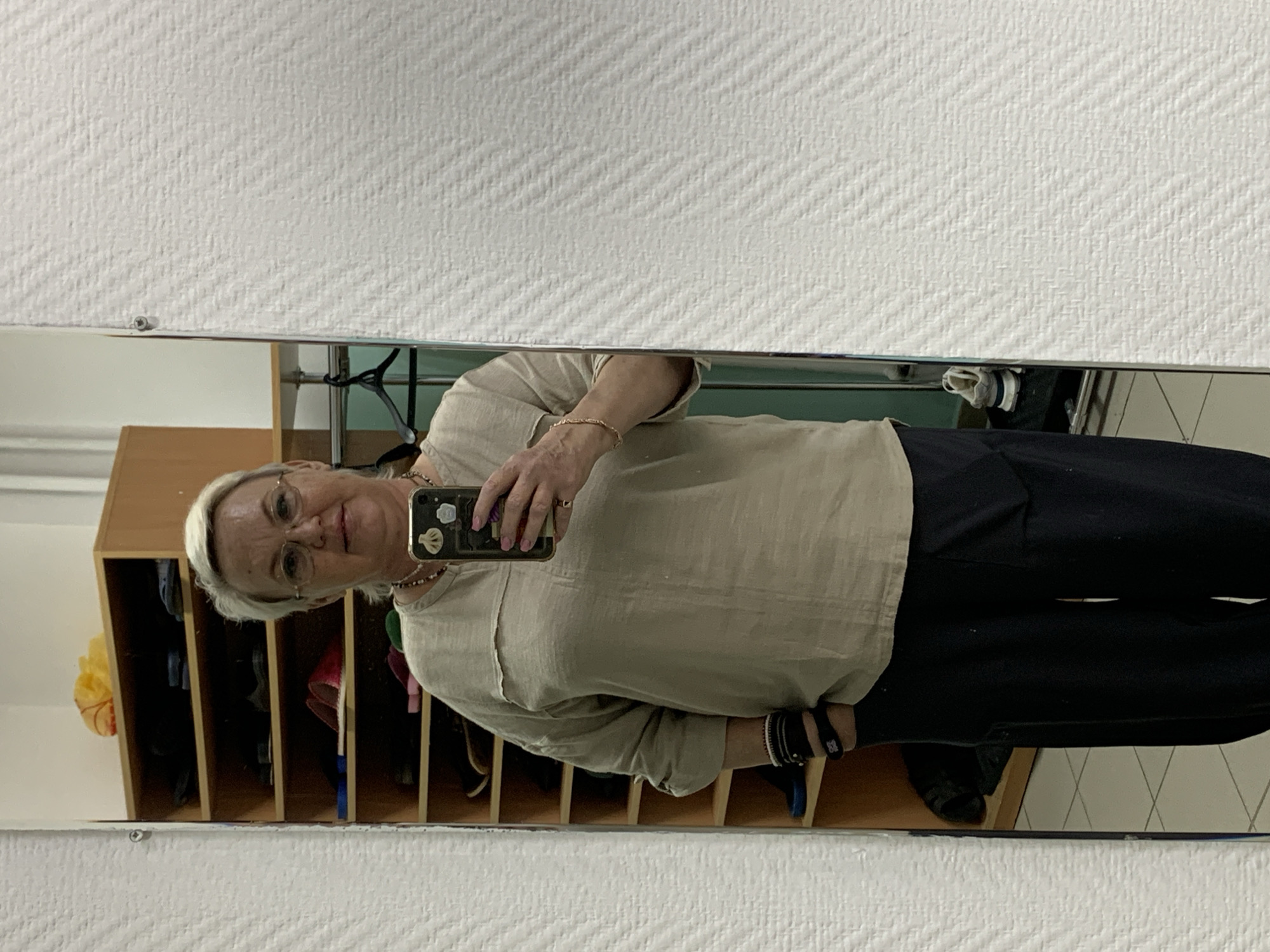 A woman in glasses and light-colored shirt with dark pants takes a mirror selfie. She is standing in front of a shelf with multiple cubbies that contain various items. The location is possibly a storage area with white-tiled walls; the city of Chaikovsky, Russia is not discernible from the image.