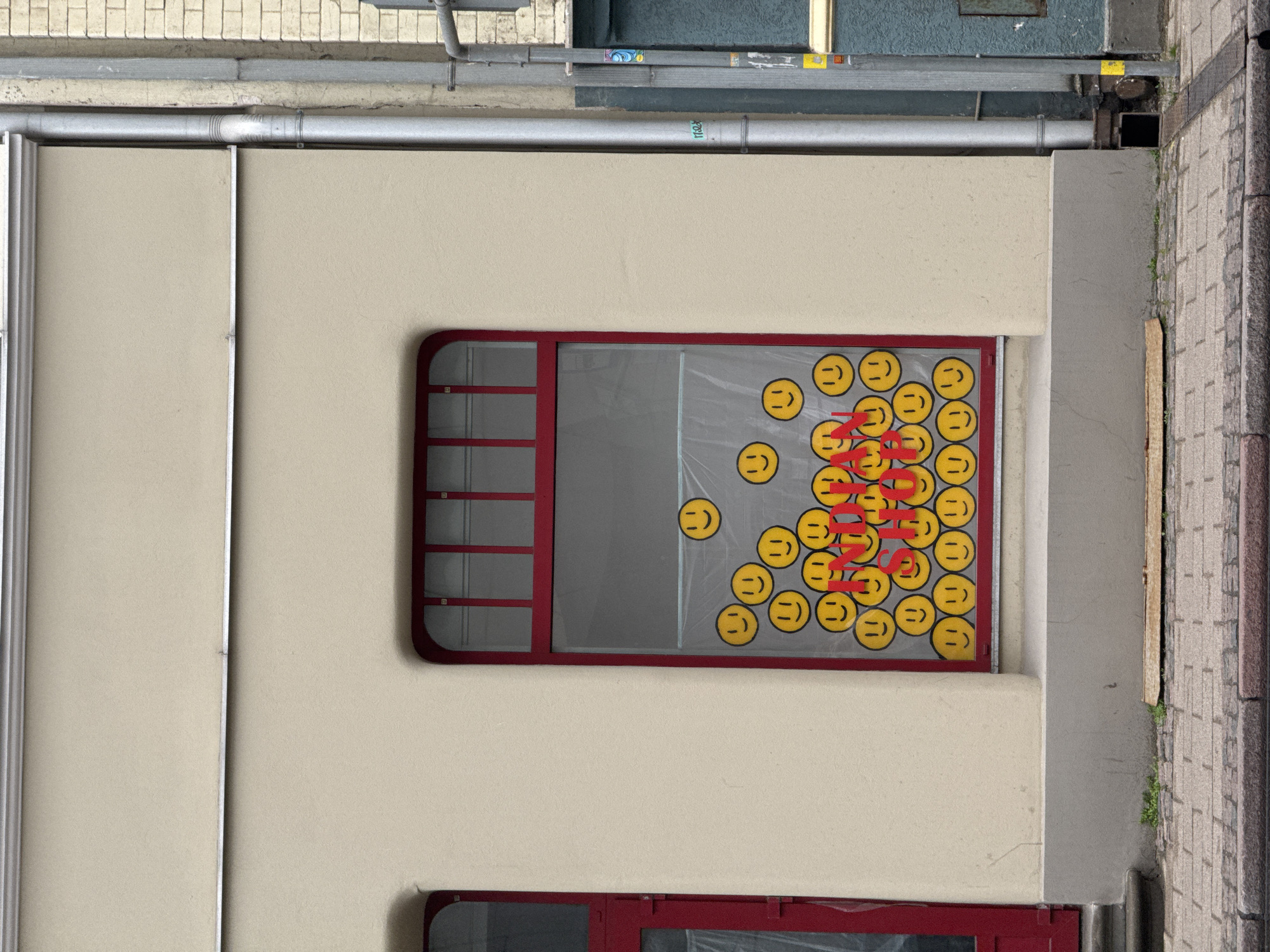 The image captures a detailed section of a building facade, presented as if viewed vertically from a street or alley. The primary subject is a cream-colored wall featuring a prominent window with a distinctive rounded-corner frame in dark red. This window has several red grilles, mainly on its left side.
Affixed to the window glass are numerous circular yellow stickers, each displaying a minimalist smiley face with two dots for eyes and a horizontal line for a mouth. These smiley faces are densely clustered towards the right side of the window. Overlapping the smiley stickers and the glass, red block letters are visible, spelling out the word "MONEY" repeatedly in at least two lines, though partially obscured by the yellow smileys.
Above the main window and along the top edge of the facade, several grey metallic pipes or conduits run horizontally. Faint text, including "MAX" and "1102X," is discernible on one of these pipes. To the right of the building wall, a cobblestone sidewalk is visible, bordered by a grey concrete-like structure. A smaller, red-framed opening is partially visible at the bottom of the image. The scene is illuminated by soft, natural light, suggesting daytime. The setting is Riga, Latvia.