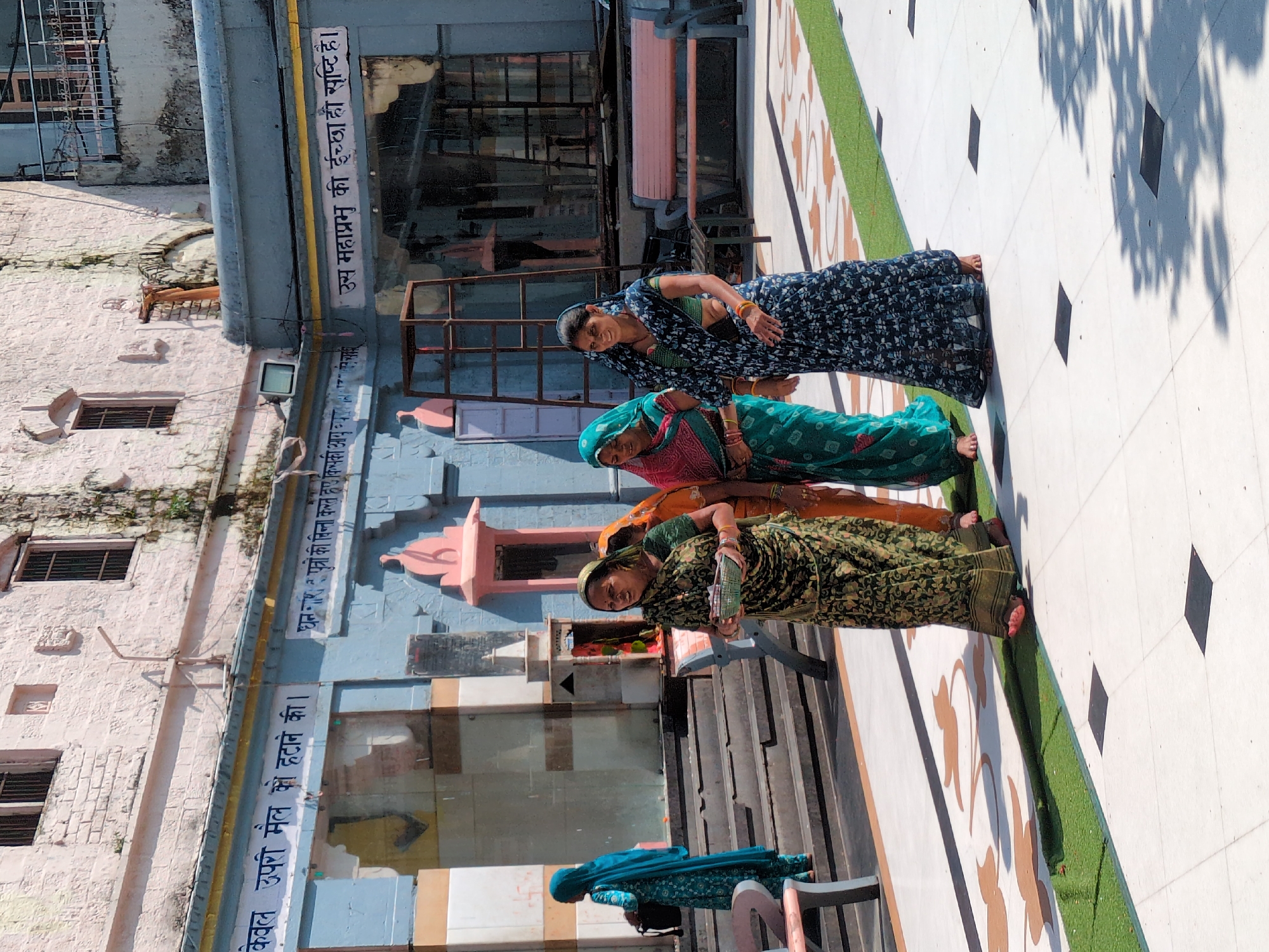 This is a medium shot photograph taken outdoors during the day, focusing on a group of four women in traditional Indian attire walking in what appears to be a temple complex or courtyard. The women are dressed in colorful sarees. The woman in the foreground, on the left, wears a dark green and black patterned saree, holding a shallow, woven green basket. Next to her, to her right, is a woman in a bright orange saree, and further right, a woman in a dark blue and white floral saree. Another woman in a turquoise blue hijab and matching patterned salwar kameez is visible in the background on the left, walking up a set of stairs.
The environment features light-colored tiled flooring with decorative brown and green patterns. In the background, a building with light pink and blue painted walls is visible, with a visible inscription in Hindi script. There are also architectural elements such as stairs, doorways, and windows with metal grills. The overall scene suggests a religious or cultural gathering. The image appears to be taken in Kangra, India, given the architectural style and the traditional Indian clothing.