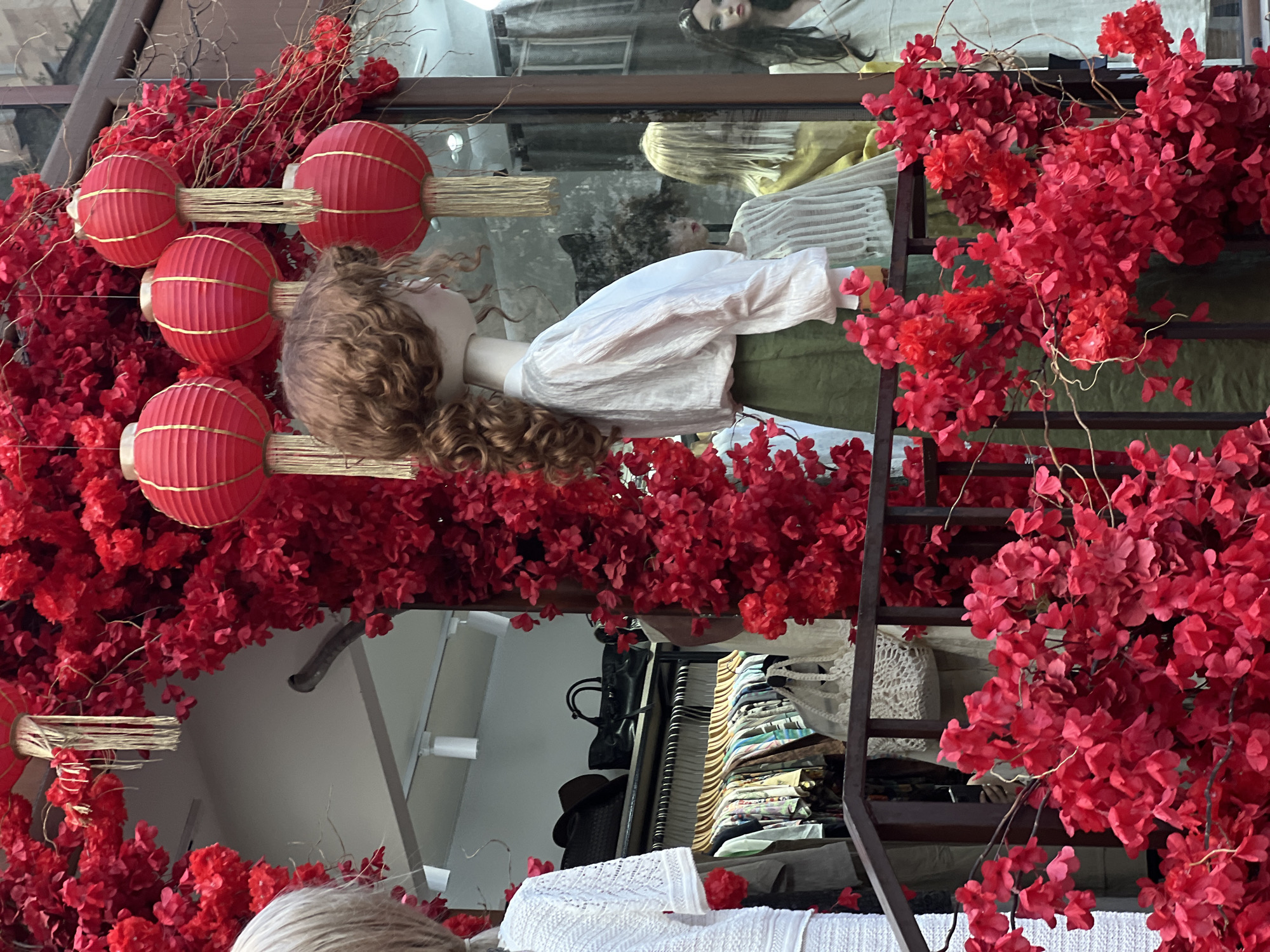 A vibrant, festive storefront display is captured, featuring a strong red theme. The central subject is a mannequin with curly brown hair, positioned on the left side of the frame, wearing a white long-sleeved top and a green skirt or trousers. Four red, accordioned paper lanterns with gold accents and wooden sticks are prominently displayed to the left of the mannequin, interspersed with dense red artificial flowers.
The entire scene is heavily adorned with these abundant red flowers, creating a lush, eye-catching border on the left, top, and bottom of the image, suggesting a lively and possibly celebratory atmosphere. Through the glass of the display window, reflections are visible: another mannequin with dark hair and what appears to be a blonde-haired mannequin are reflected higher up and behind the central figure, respectively. Further reflections reveal elements of the shop's interior, including clothing racks filled with garments, hats, and bags. A dark metal railing or ladder-like structure is also visible through the red flowers on the right side. At the bottom right, a white knitted or crocheted item is part of the display.
The setting is clearly a retail boutique, likely showcasing clothing. The combination of red lanterns and copious red blossoms suggests a festive decoration, possibly for an event like Lunar New Year, Valentine's Day, or a general vibrant seasonal display. The lighting appears bright, indicative of daytime. There is no visible text in the image, and no specific activity is taking place beyond the static display.